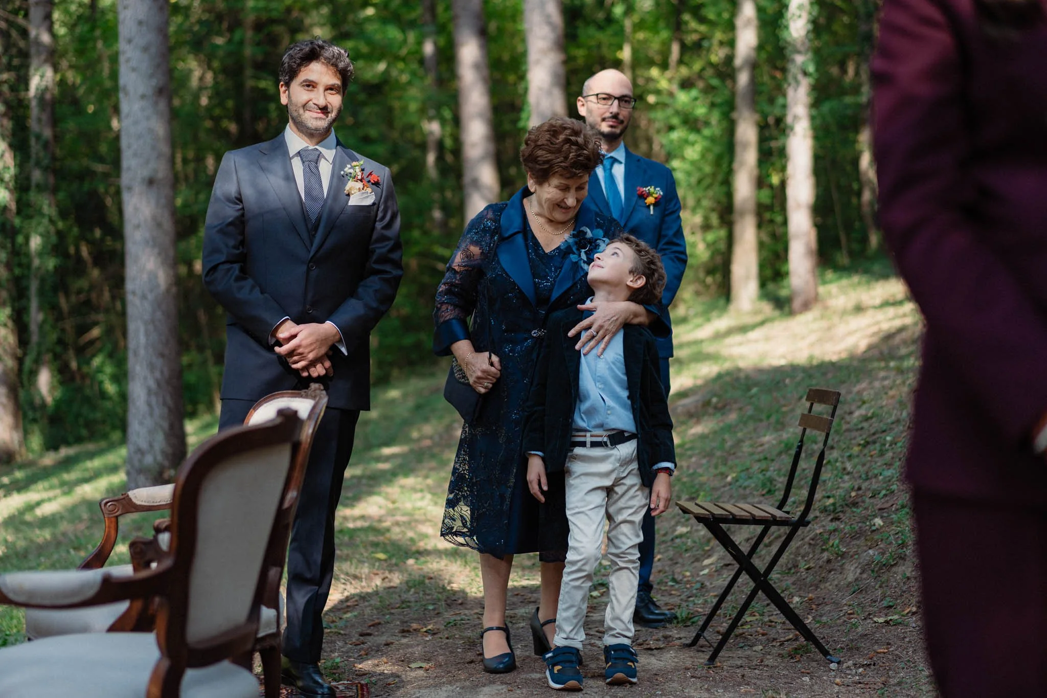 Gruppo di persone alle nozze in un bosco, uomini in abiti eleganti, donna e bambino in abiti formali, tutti sorridenti.