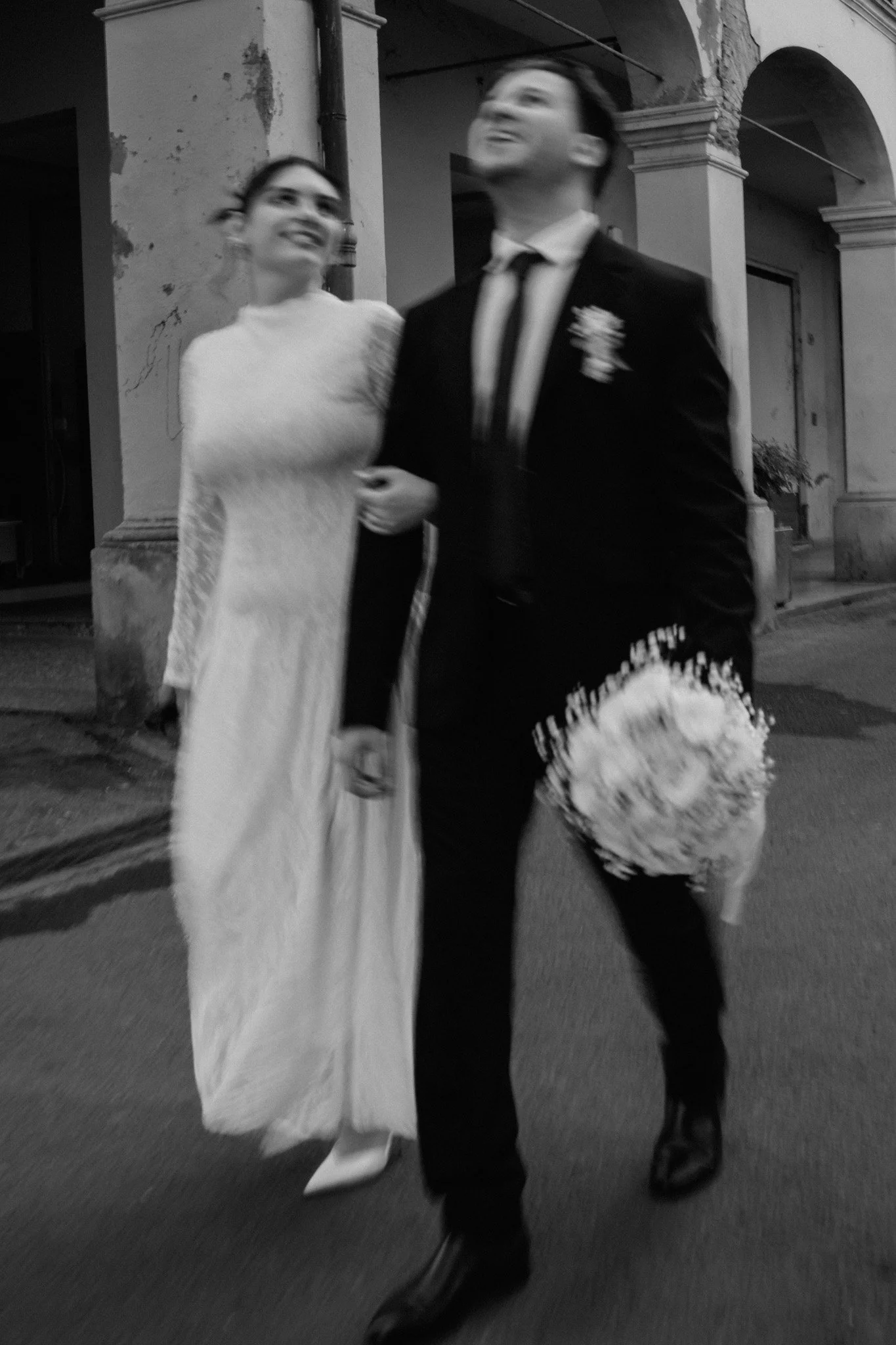 Due persone, una donna in abito lungo bianco e un uomo in completo nero, camminano insieme in strada, con l'uomo che tiene un bouquet di fiori.