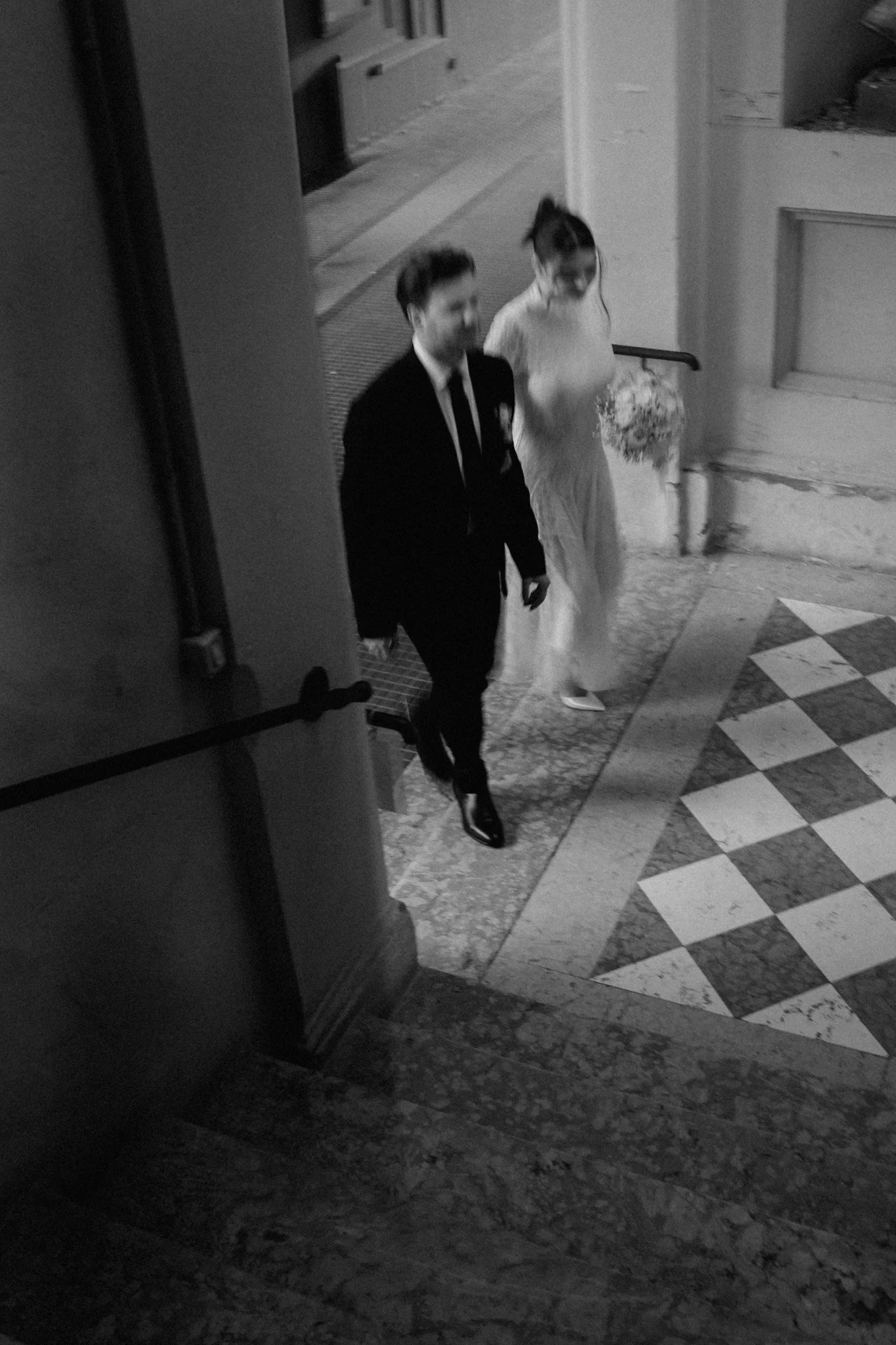 Due persone vestite elegantemente, uno uomo in abito nero e una donna in abito chiaro, che camminano su un ingresso in un edificio.