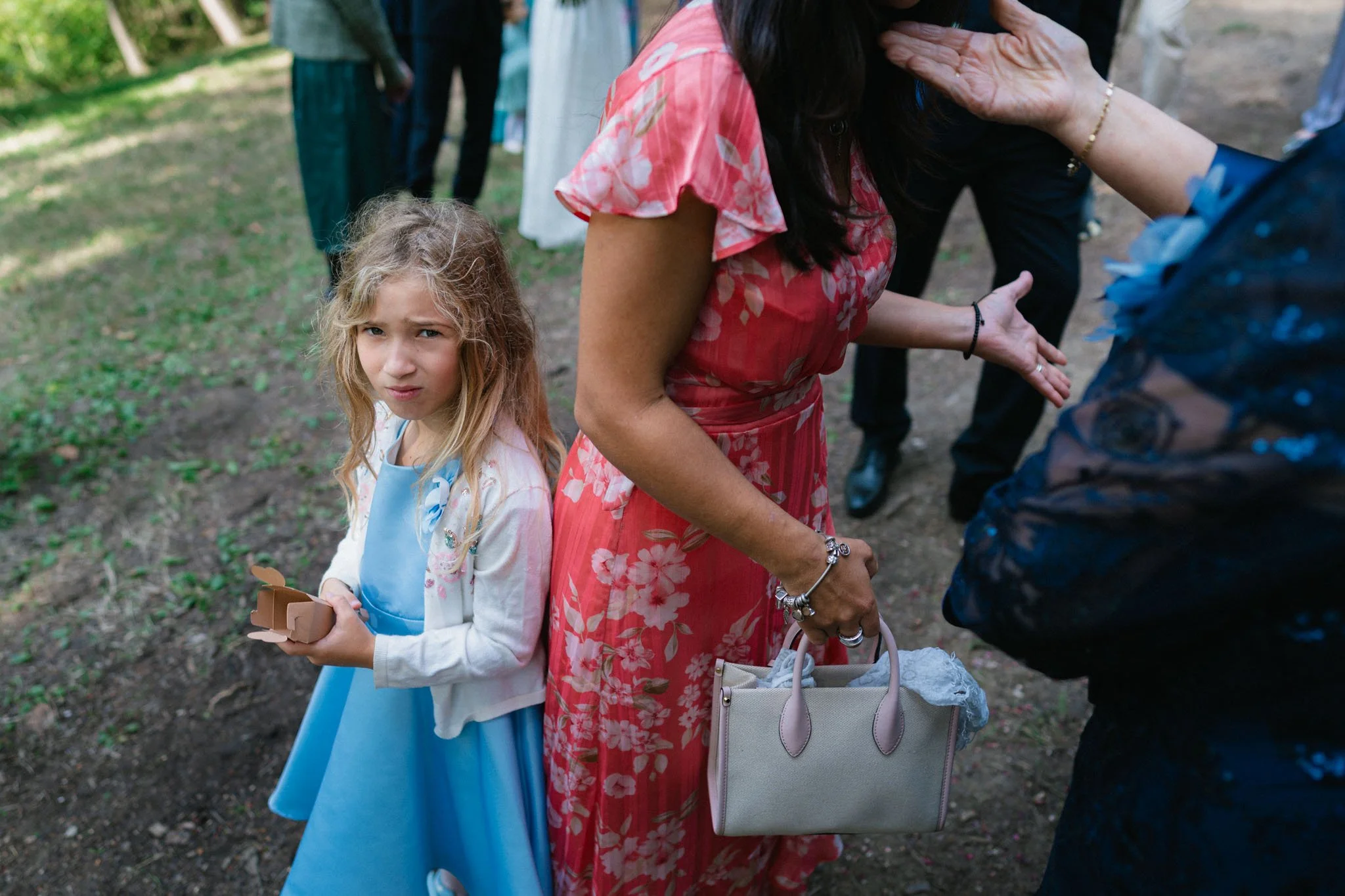 Una bambina vestita con un abito azzurro e una giacca bianca tiene una scatola in mano e guarda con un'espressione perplessa. Dietro di lei, una donna con un vestito a fiori rossi e una borsa beige sta interagendo con una persona non visibile. L'imma