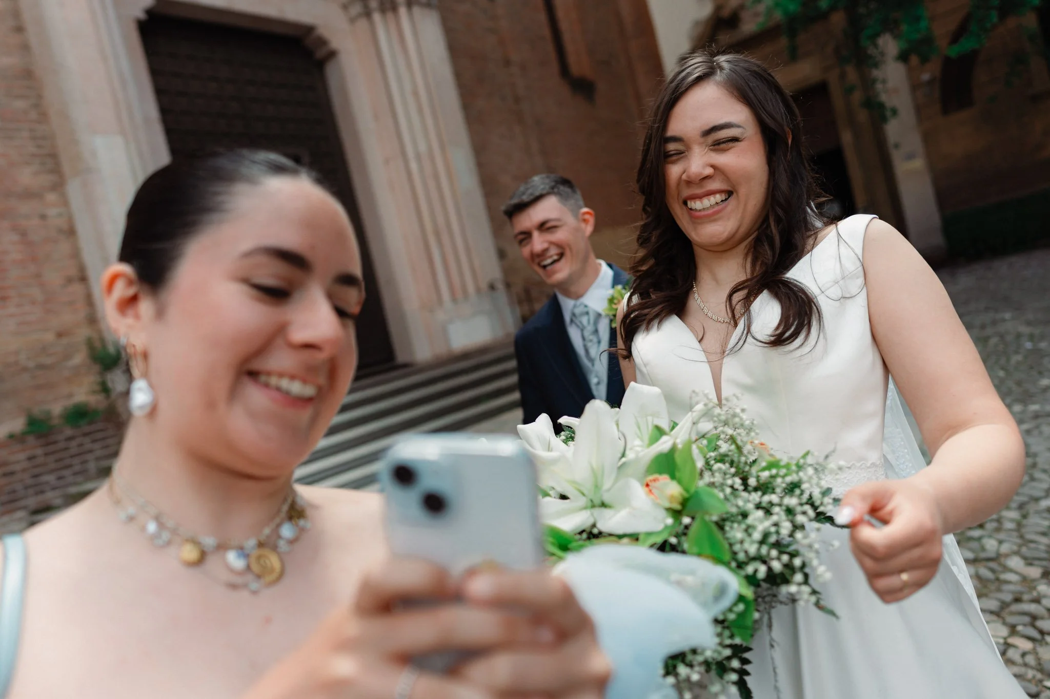 Tre persone sorridenti che scattano una foto con uno smartphone durante un matrimonio, con un edifico storico sullo sfondo.