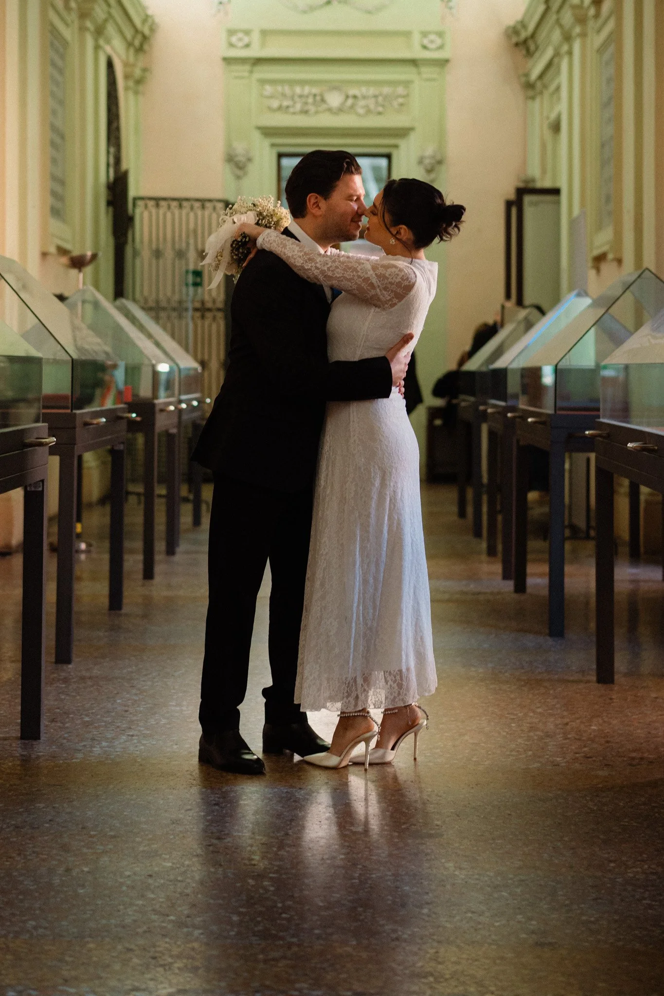 Una coppia di sposi si abbraccia e si bacia nel giorno del loro matrimonio in una sala decorata con dettagli antichi, davanti a vetrine di esposizione.