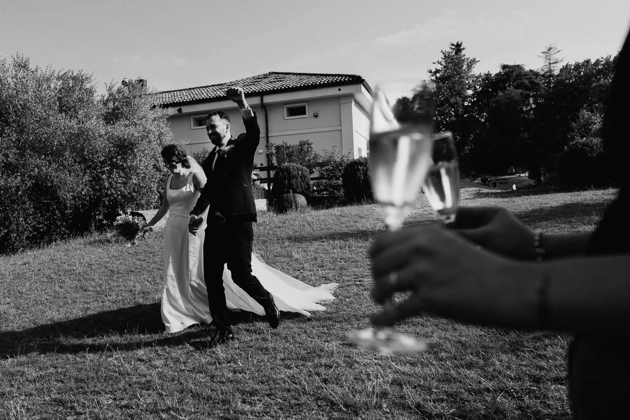 Sposi di sposi che si tengono per mano e festeggiano sulla scena, con una mano che tiene un bicchiere di vino in primo piano, in un ambiente esterno verde e soleggiato.
