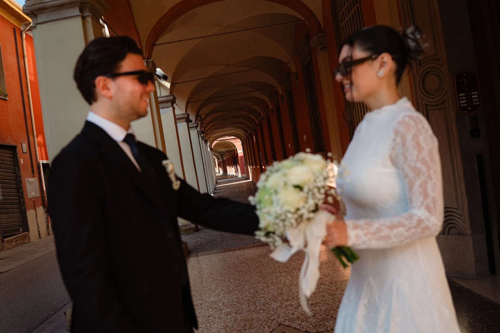 Una coppia di sposi sorridenti si scambia un gesto romantico in un arco di un edificio storico, l'uomo indossa un abito da uomo e la donna un vestito da sposa bianco. Sono entrambi con occhiali scuri, l'uomo tiene un bouquet di fiori e la scena è ill