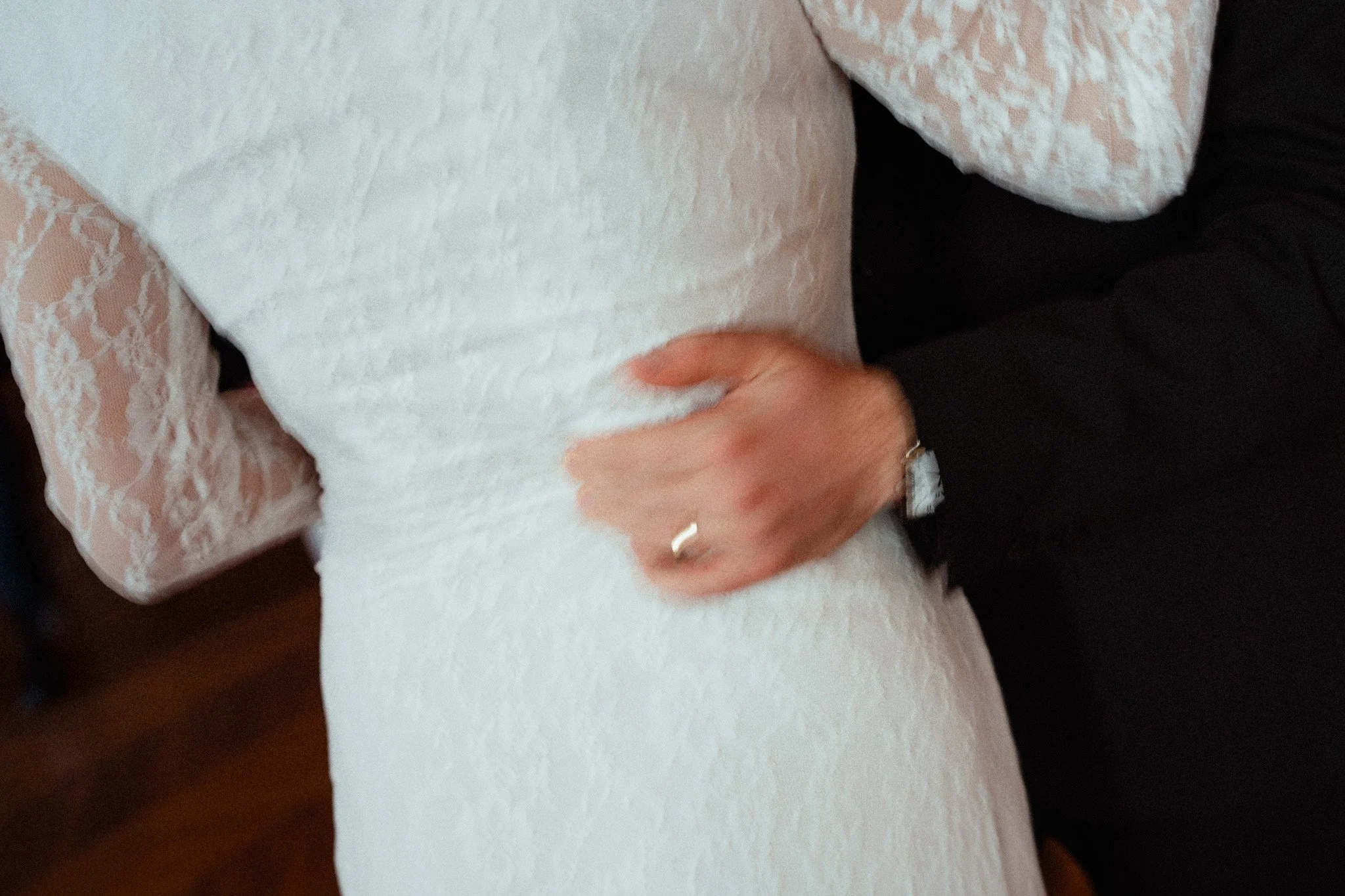 Due persone abbracciate, una con un abito bianco in pizzo e l'altra con un abito nero, si tengono stretti in un gesto affettuoso.