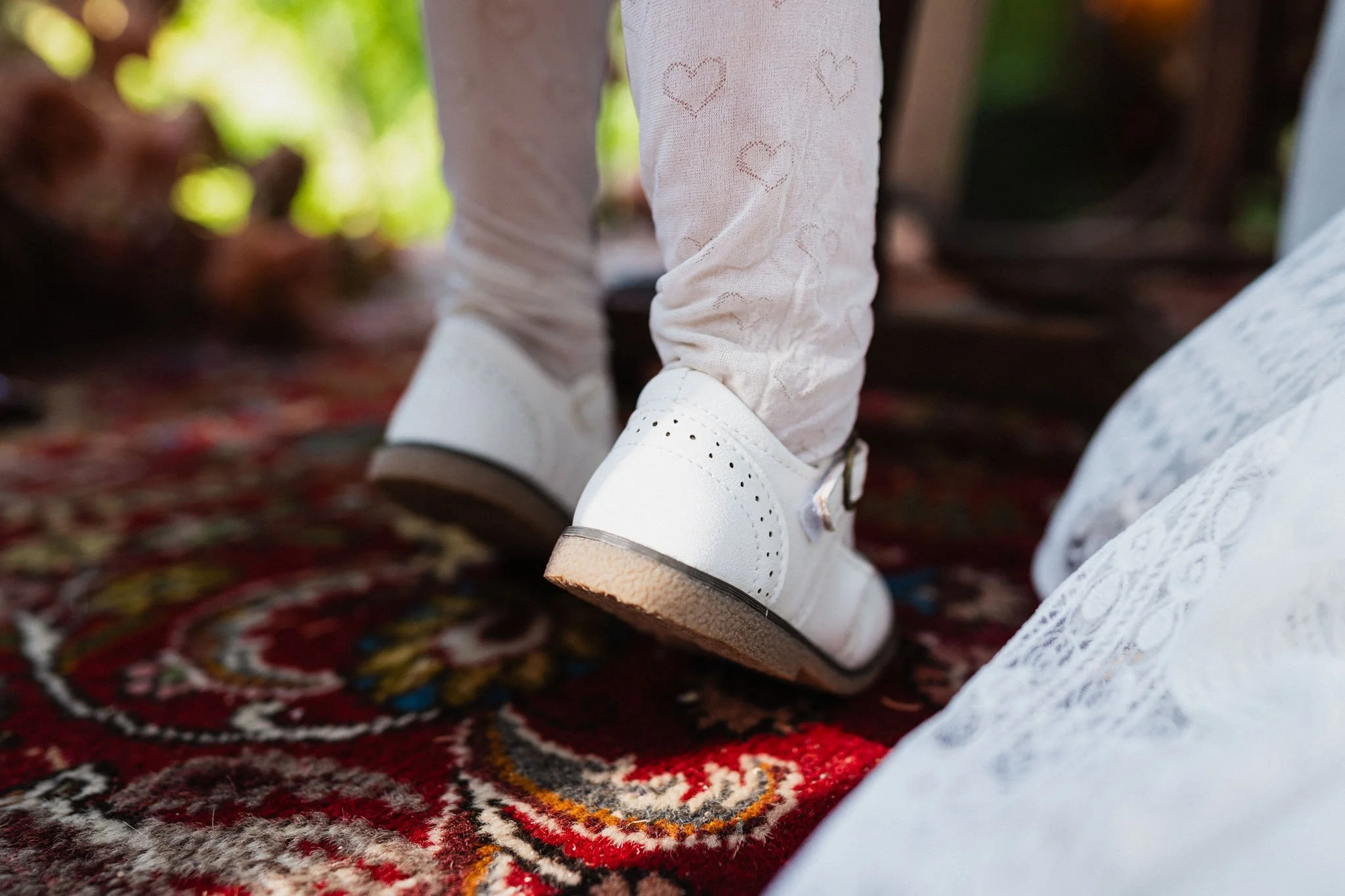 Piedi di una persona con scarpe bianche, pantaloni beige con disegni di cuori, su un tappeto rosso con motivi decorativi.