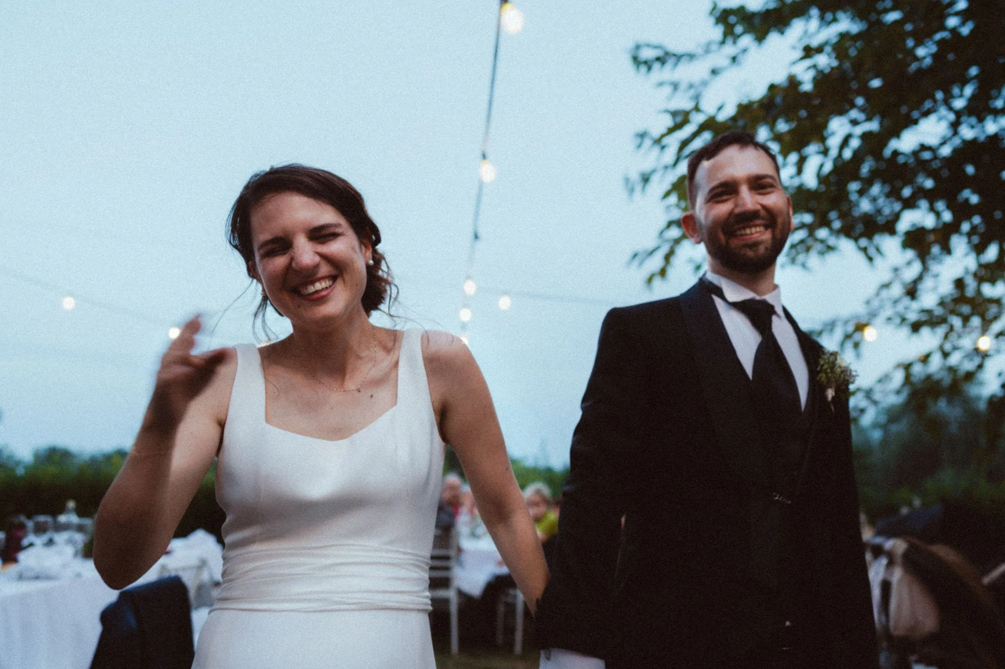 Una coppia di sposi felici durante una cerimonia all'aperto in serata, con luci decorative e persone sullo sfondo.