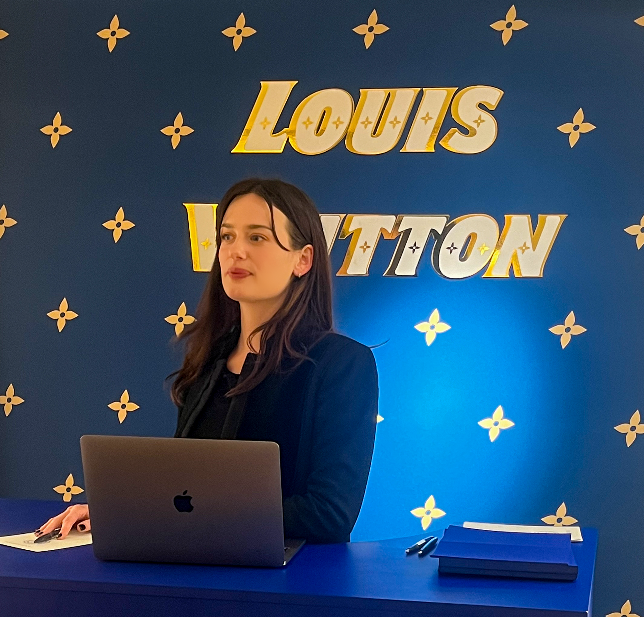 Une femme avec de longs cheveux foncés se tient derrière un bureau bleu, devant un fond décoratif avec le texte "Louis Vuitton" en lettres dorées. Elle a un ordinateur portable Apple et un bloc-notes sur le bureau.