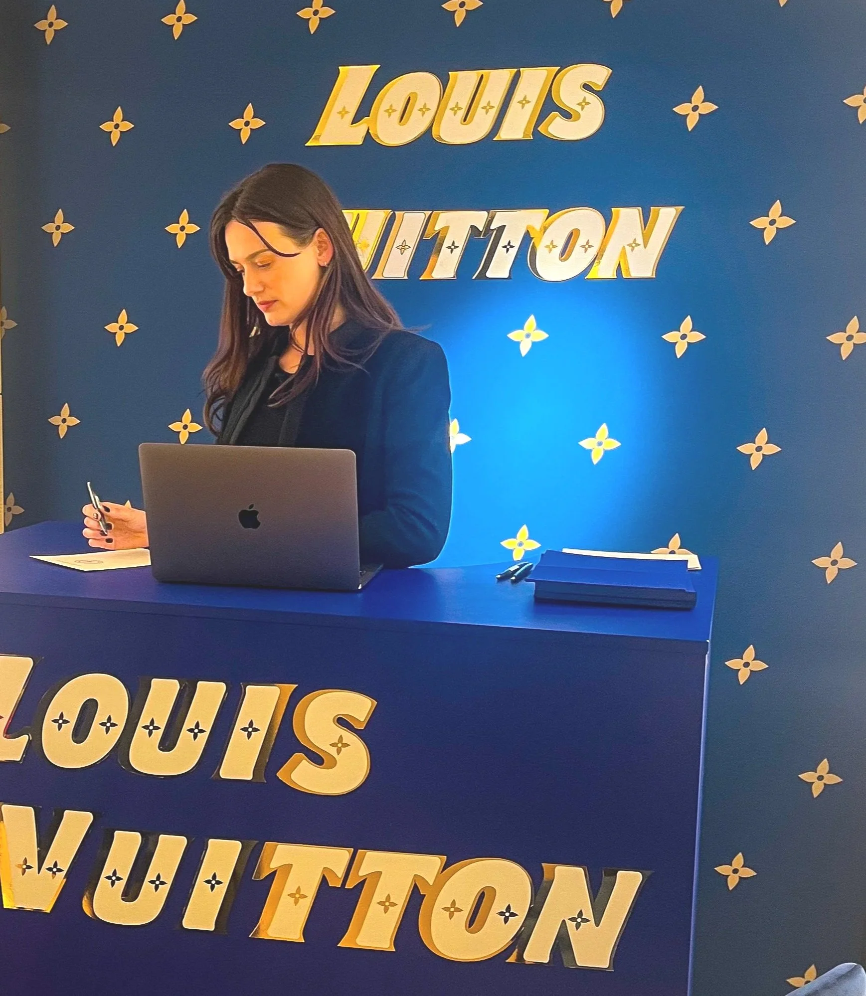 Séverine (Chandrastrology) derrière un comptoir de luxe Louis Vuitton, avec un mur bleu en arrière-plan portant le logo Louis Vuitton en lettres dorées.