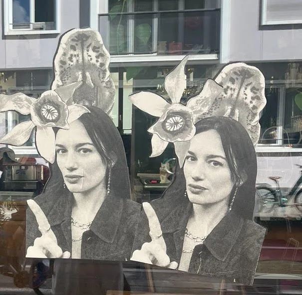Deux affiches en noir et blanc de la même femme, décorées avec des fleurs et des feuilles en papier, placées derrière une vitre d'une vitrine.