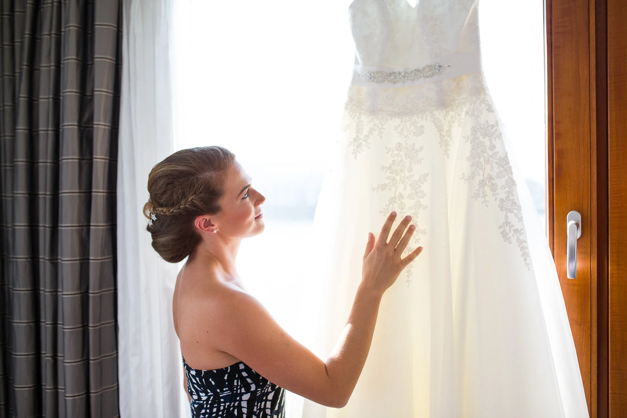 Eine Frau mit hochgestecktem Haar betrachtet ein weißes Hochzeitskleid, das an einem Fenster hängt.