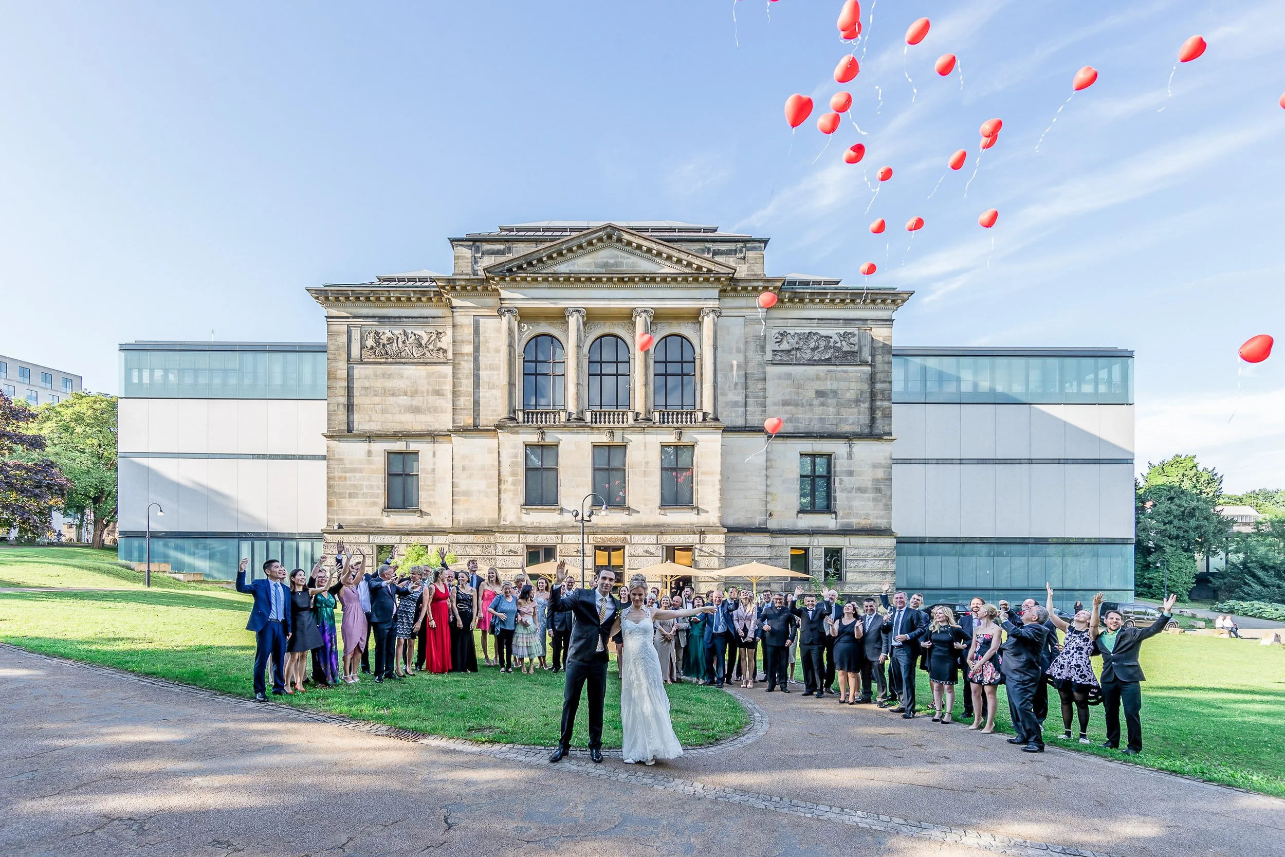 Hochzeitspaar vor einem historischen Gebäude, umgeben von Gästen, bei einer festlichen Feier, mit roten Ballons am Himmel