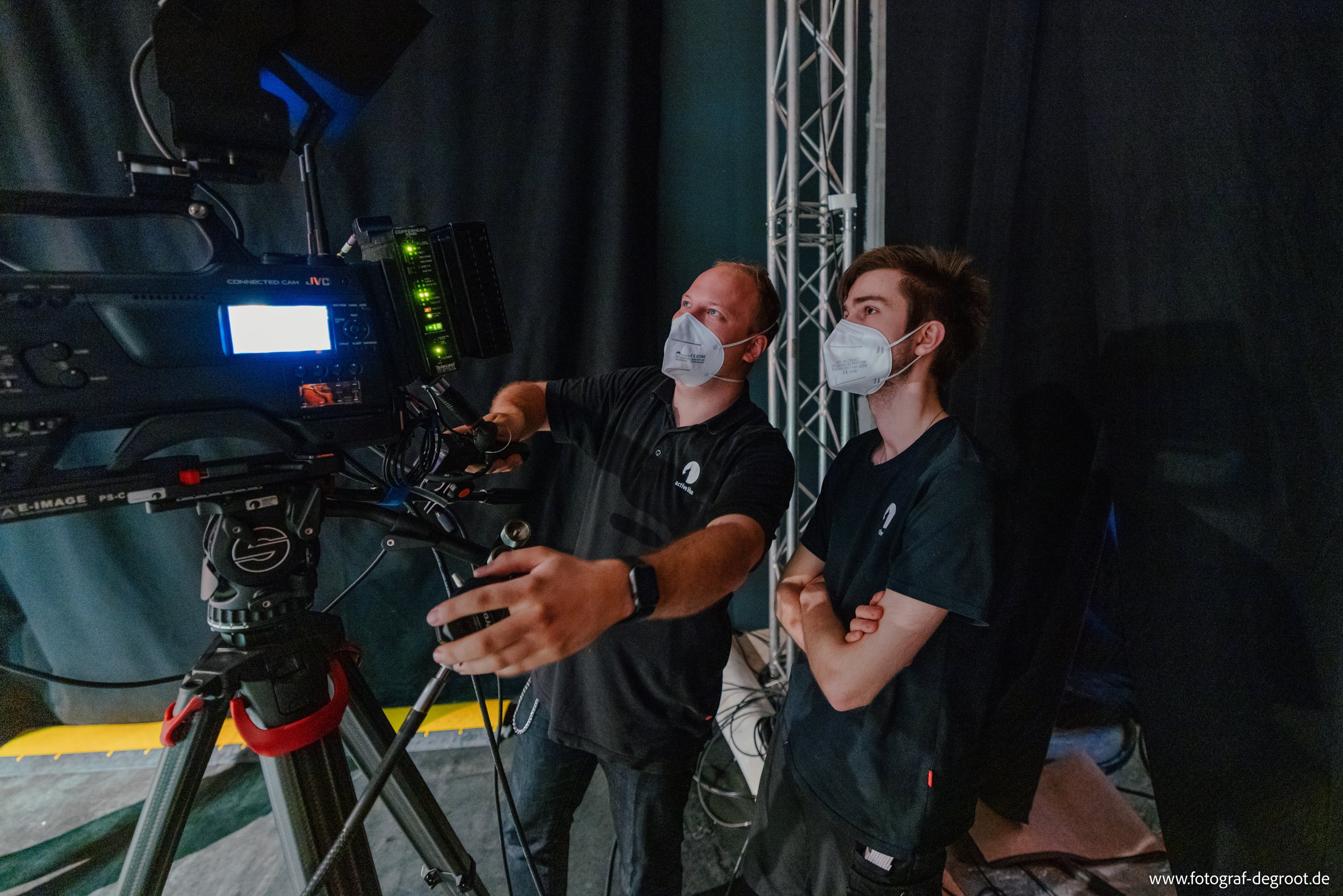 Zwei Männer tragen Masken und stehen neben einer professionellen Videokamera. Sie befinden sich in einem dunklen Raum mit Vorhängen. Einer der Männer zeigt auf die Kamera, als ob er etwas erklärt. Es gibt technische Ausrüstung im Hintergrund, einschl