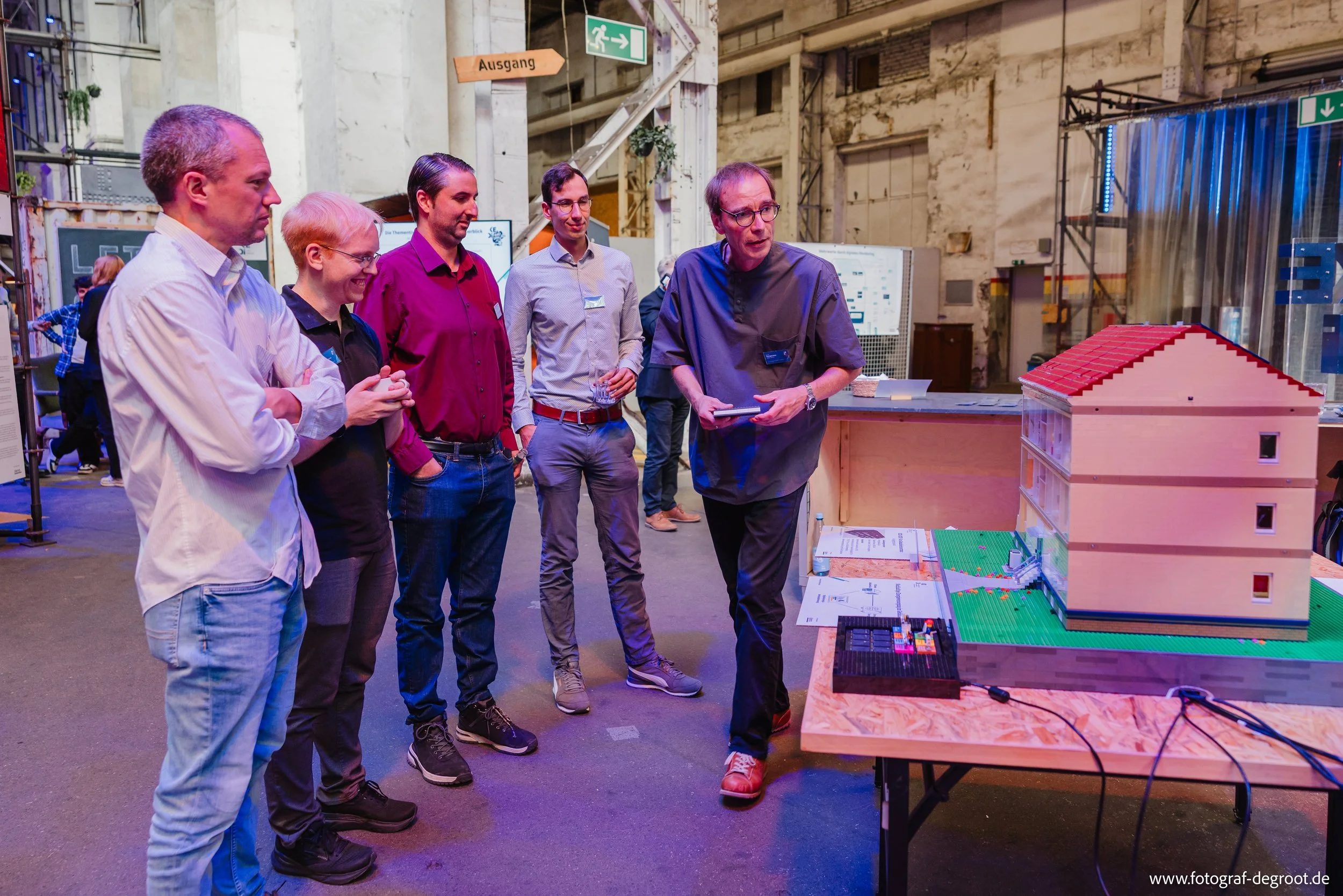 Eine Gruppe von fünf Menschen, die einem Mann zuhören, der eine Präsentation hält, während er auf eine Miniatur eines Hauses aus LEGO-Steinen zeigt. Die Szene findet in einem industriellen Ambiente statt, wahrscheinlich auf einer Messe oder Ausstellu