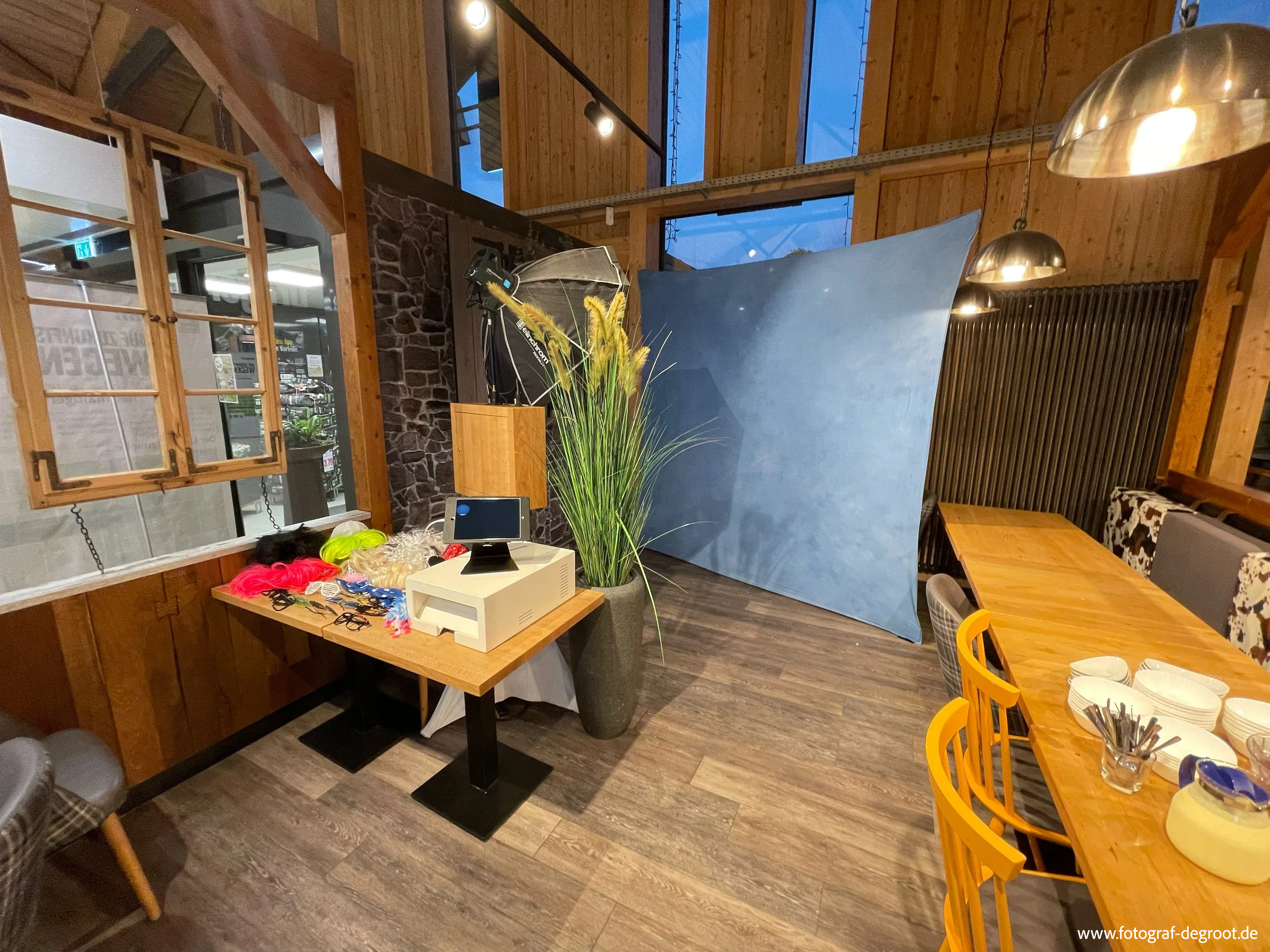 Innenansicht eines Raumes mit Holzdecke und Holzverkleidung, ein Tisch mit weißen Schalen, Stühlen in Gelb, ein großer Topf mit Pflanze, Studioausrüstung mit Hintergrund-Projektor, Fenster, und verschiedene Gegenstände auf einem kleinen Tisch.