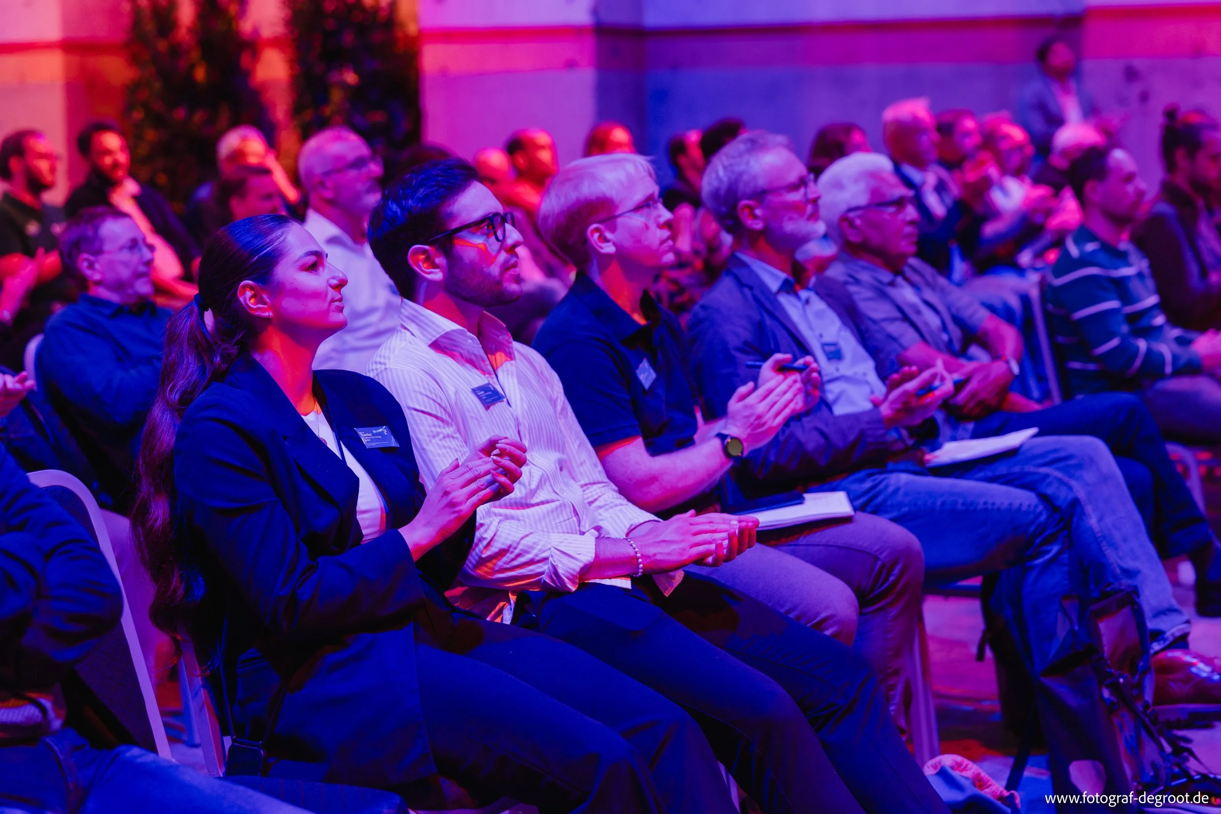 Menschen sitzen in einem Auditorium bei einer Konferenz oder einem Vortrag, beleuchtet mit buntem, vor allem pinkem Licht, während sie zusehen und zuhören.