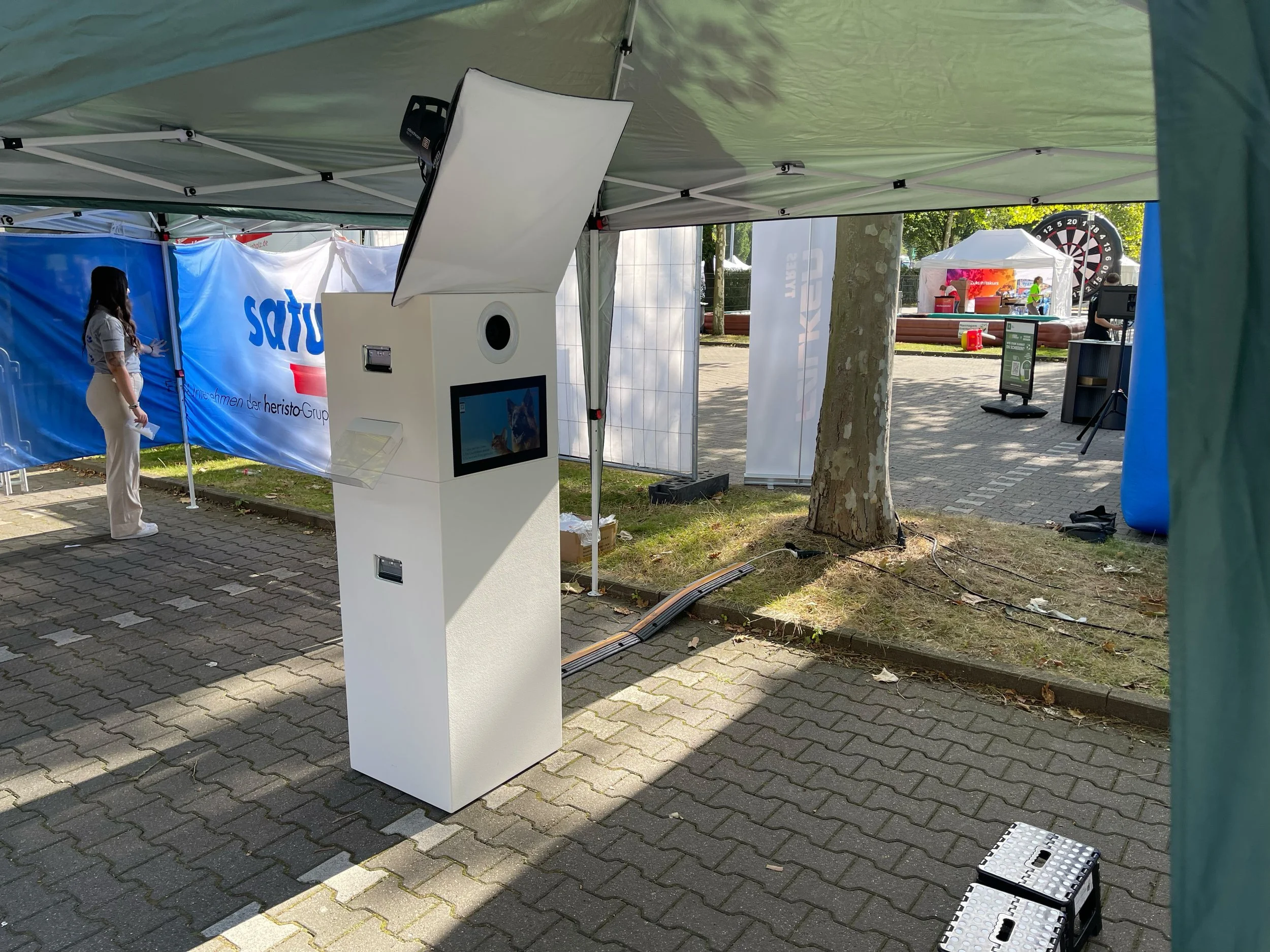 Ein Foto von einem Bewegungs- oder Fotokiosk im Freien, umgeben von Zelten und Menschen, vermutlich bei einer Veranstaltung oder Messe, mit einem Baum im Hintergrund und einem Spielautomaten oder Zielscheibe in der Ferne.