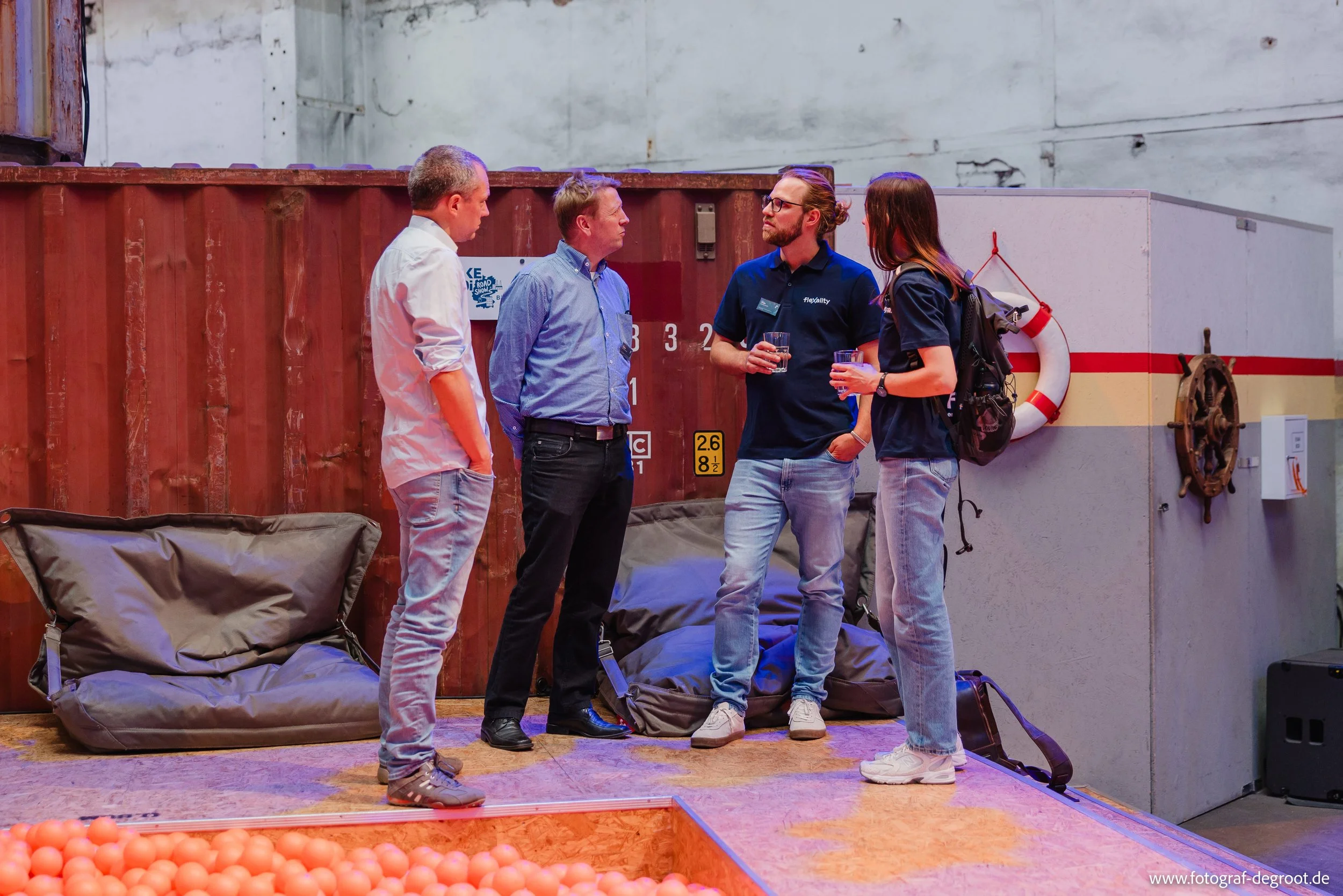 Vier Menschen stehen im Gespräch in einem Indoor-Spielplatz mit Ballgrube. Im Hintergrund eine rote Containerwand und eine weiße Wand mit einem Rettungsring und einem Steuerrad.