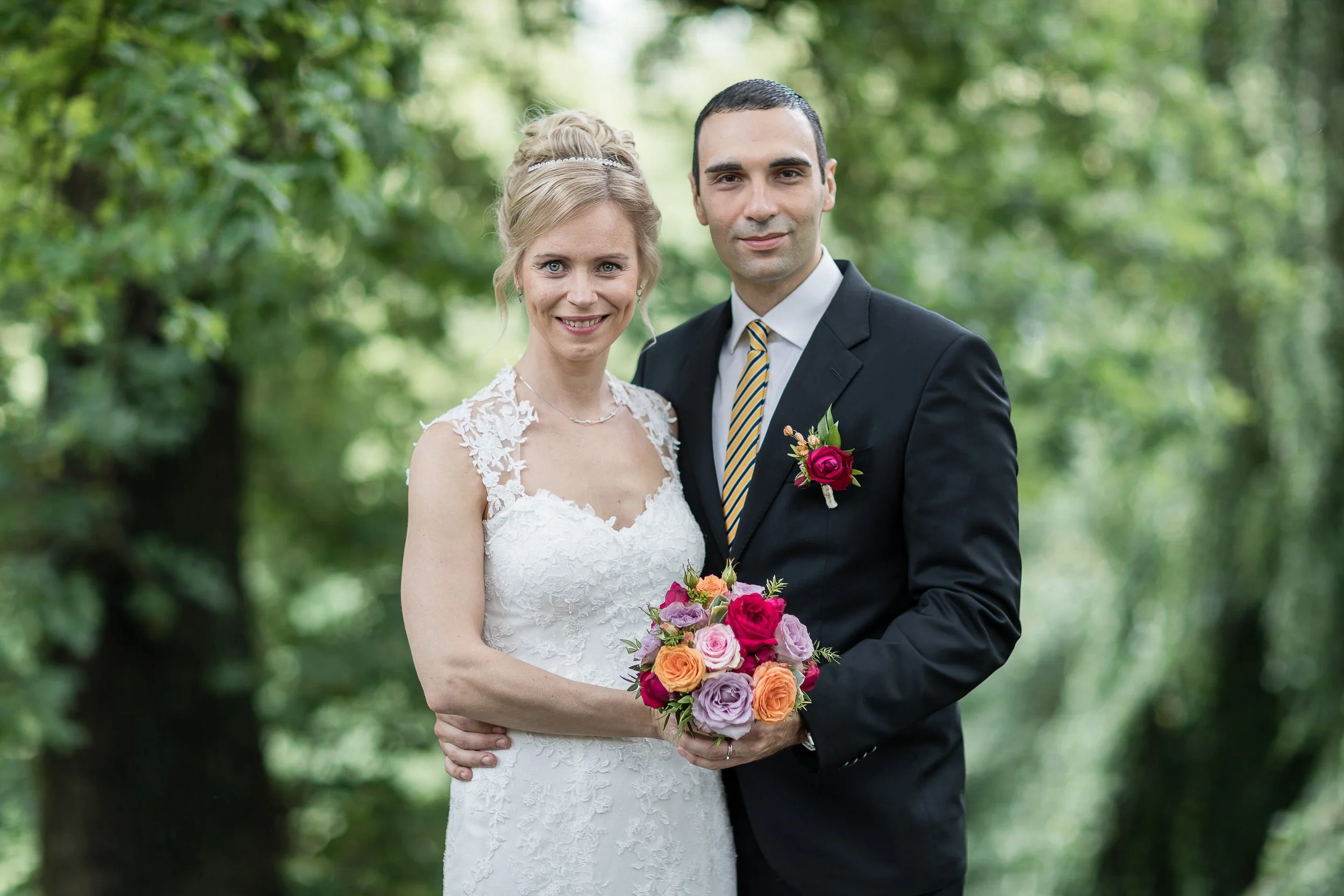 Braut und Bräutigam stehen im Wald, die Braut trägt ein weißes Hochzeitskleid mit Spitzen-Details und hält einen bunten Blumenstrauß, der Bräutigam trägt einen schwarzen Anzug mit gelb-blau gestreiftem Krawatte.