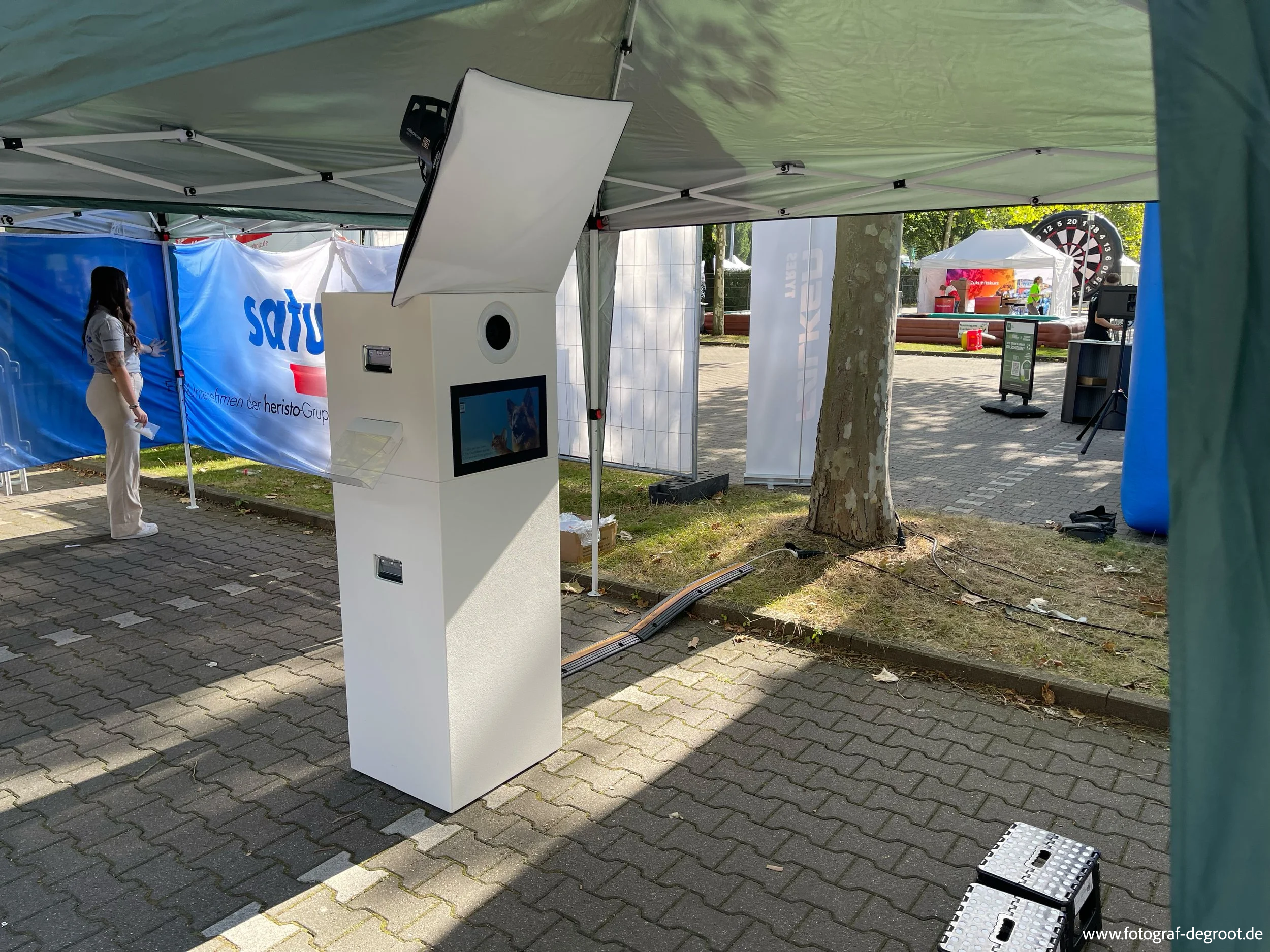 Ein fotobooth mit Monitor und Drucker in einem Zelt bei einer Veranstaltung im Freien, im Hintergrund sind Menschen, ein Dart-Spiel und Stände sichtbar.