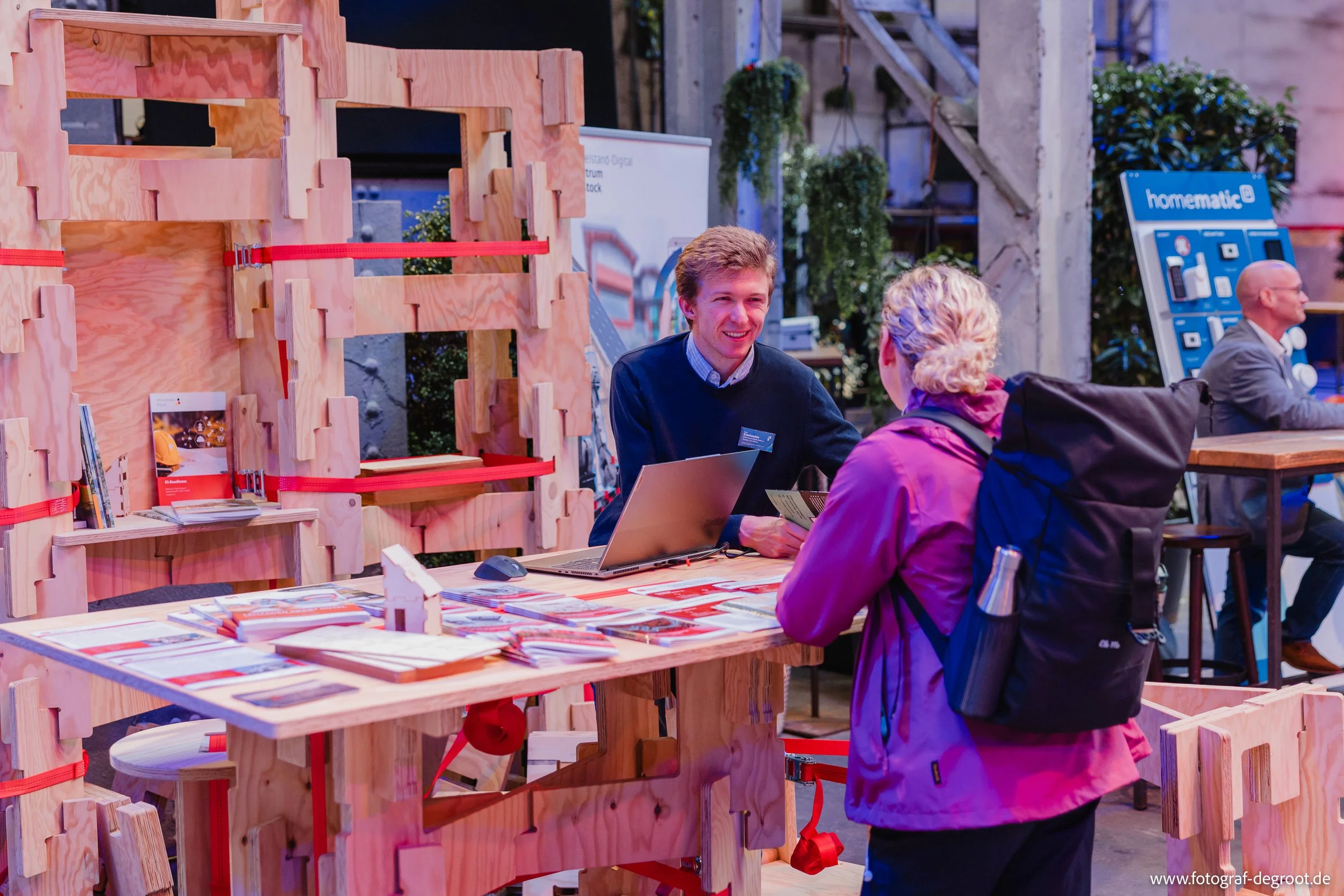 Zwei Personen sprechen an einem Holz-Freistand bei einer Messe oder Ausstellung, hinter ihnen weitere Aussteller. Der Mann hinter dem Stand lächelt und trägt eine dunkle Jacke, die Frau trägt eine pinke Jacke und einen schwarzen Rucksack.
