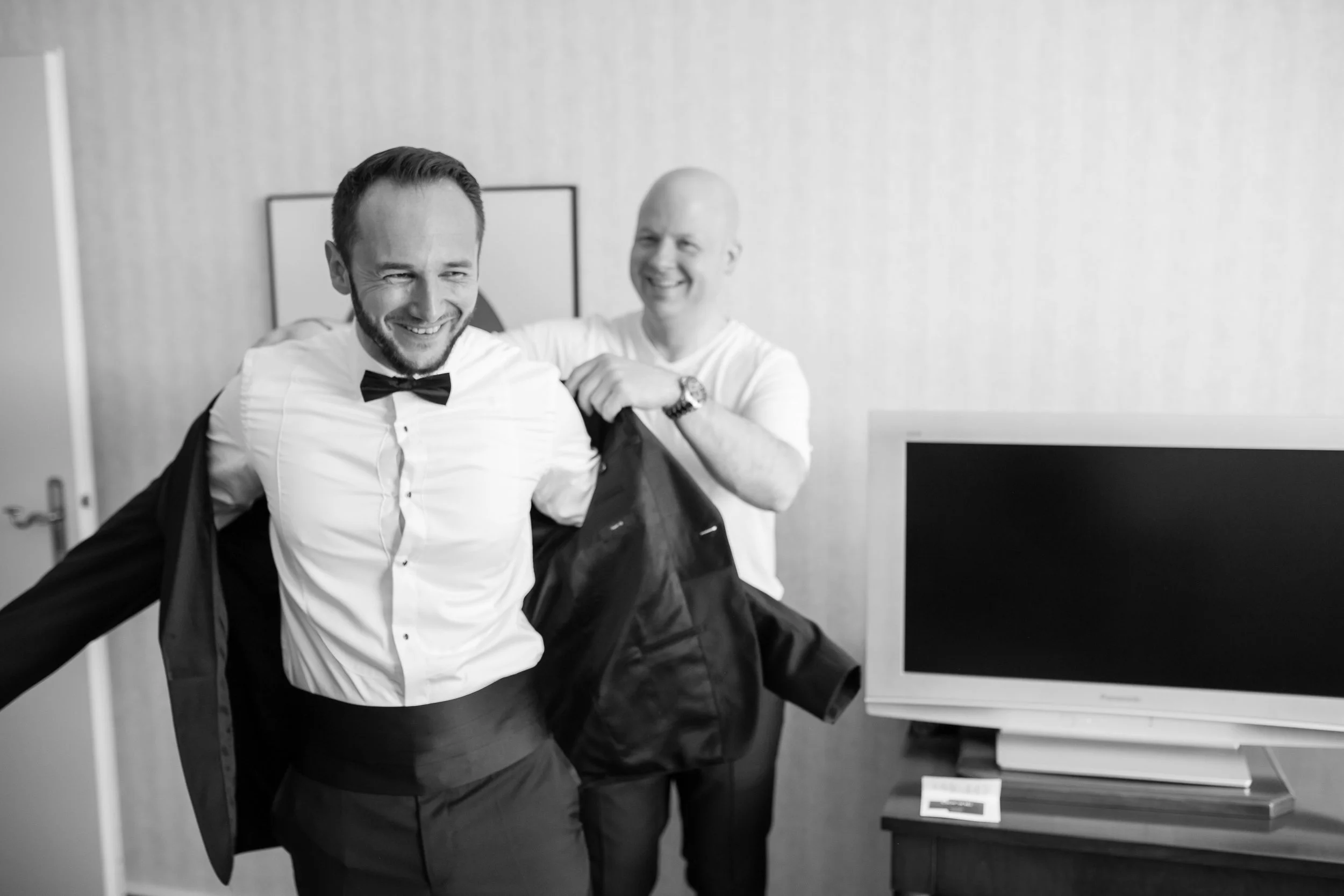 Zwei Männer in formeller Kleidung lachen in einem Raum, wobei einer den anderen während des Anziehens eines Anzugs hilft.