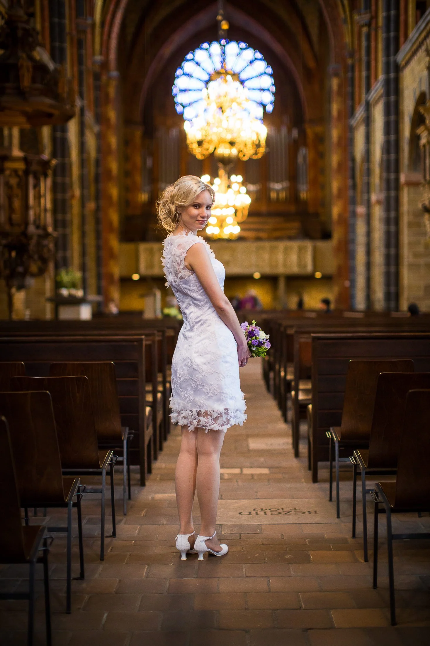 Eine Frau in einem weißen Hochzeitskleid mit Blumenmuster steht in einer Kirche mit Holzbänken und buntem Glasfenster im Hintergrund, hält Blumenstrauß in der Hand, trägt weiße High Heels und schaut in die Kamera.
