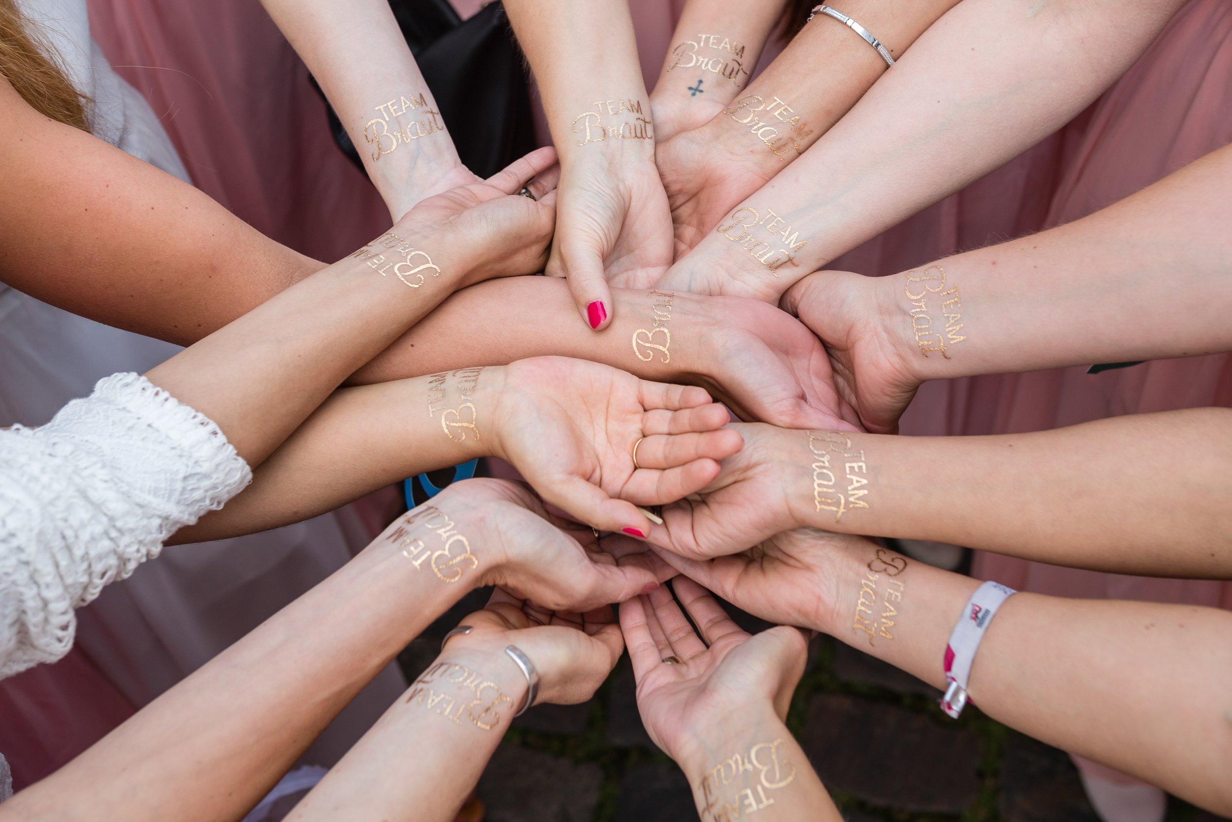 Mehrere Frauen legen ihre Hände übereinander, alle mit Tattoos, die 'TEAM Braut' in gold Schrift anzeigen.