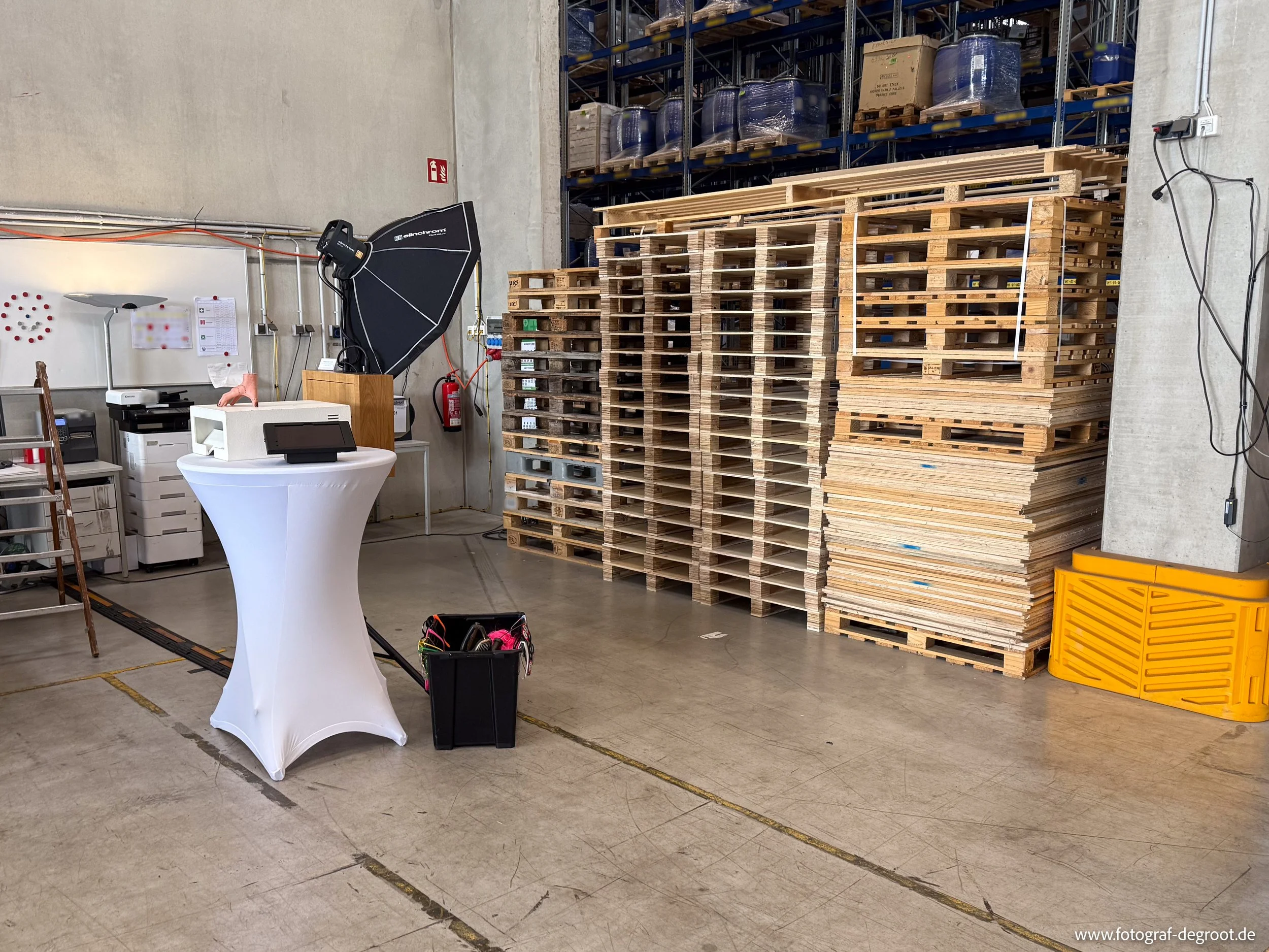 Lagerraum mit Stapeln aus Holzpaletten, Fotostudio mit Kamera-Equipment und einem Stehtisch im Vordergrund.
