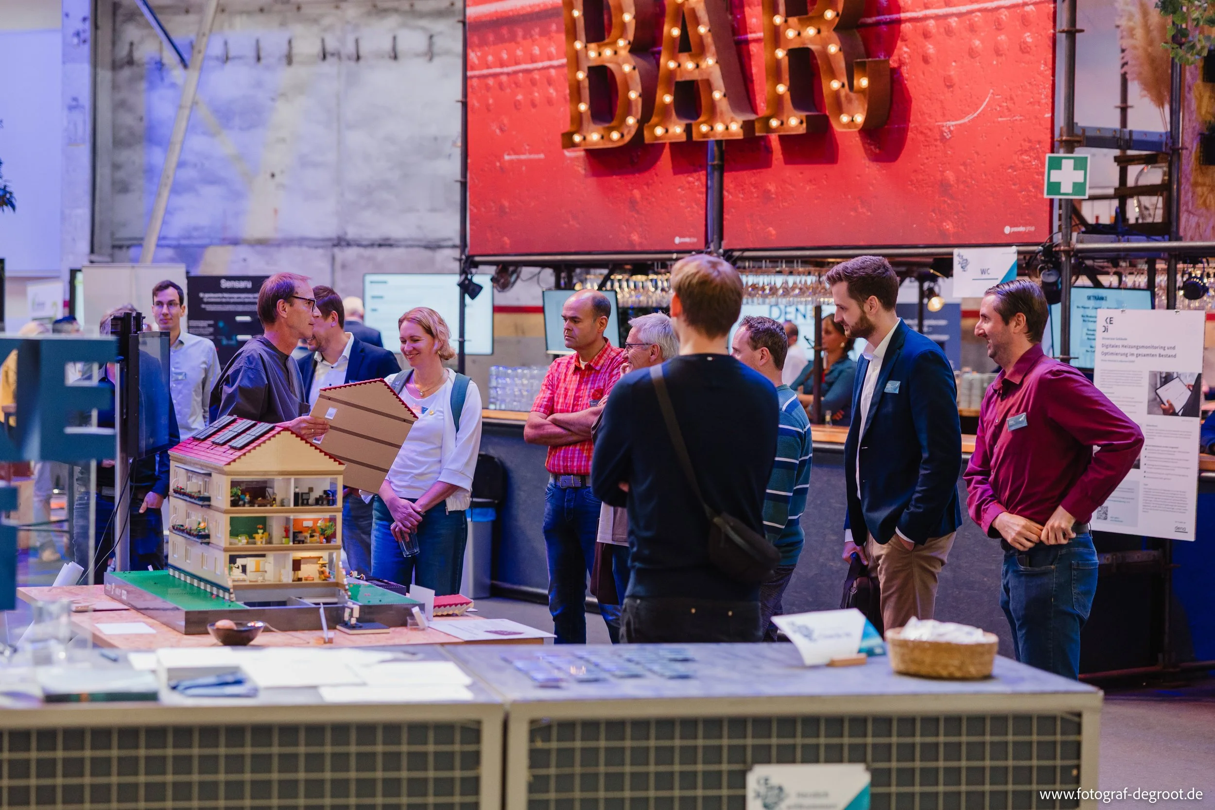 Menschen besuchen einen Marktstand mit Miniatur-Modell eines Hauses, im Hintergrund ein großes rotes Schild mit bunten Buchstaben und Lichter. Es gibt mehrere Personen im Gespräch, einige tragen Namensschilder.