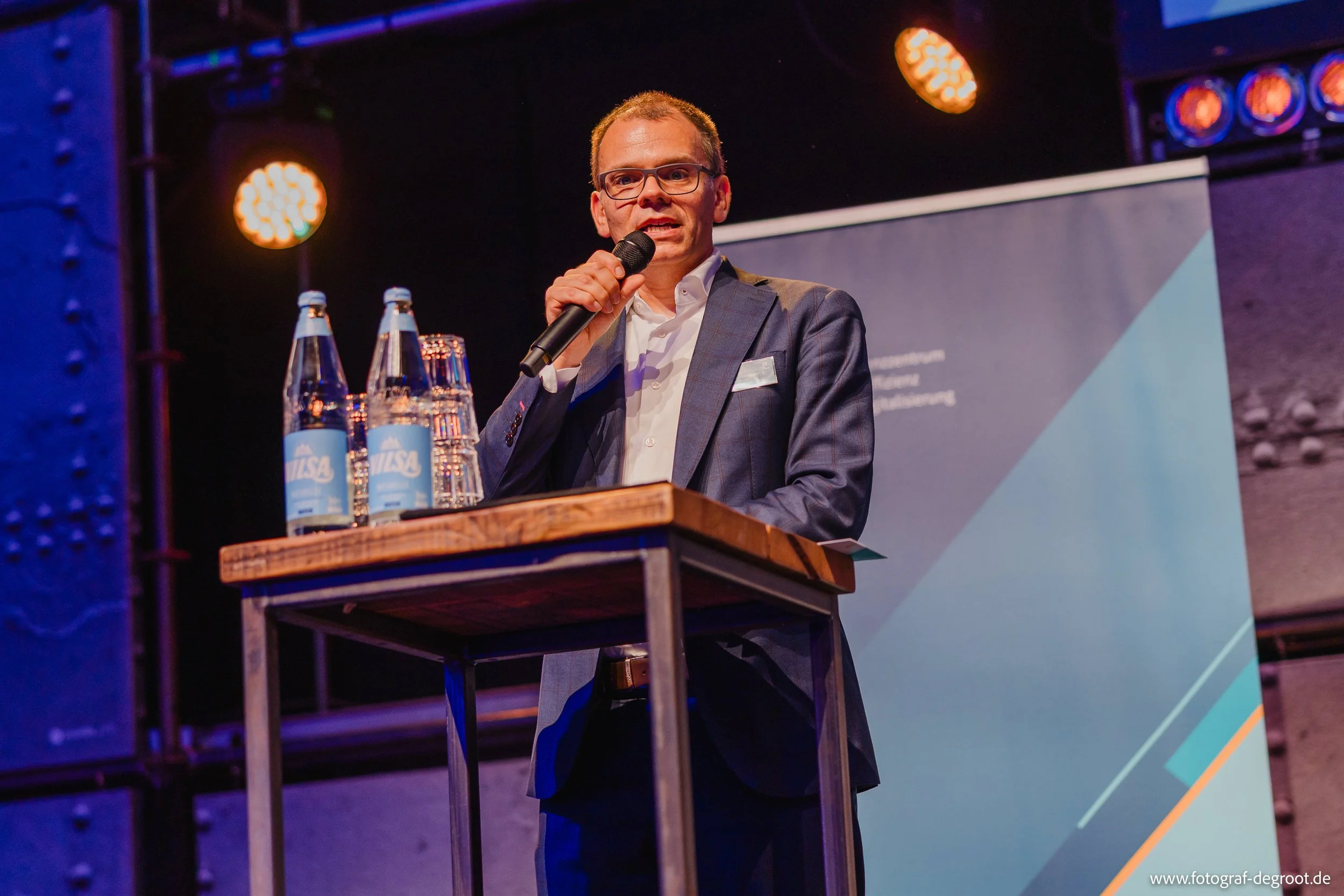 Mann in Anzug hält Mikrofon bei Vortrag auf Podium, im Hintergrund Banner, auf Tisch Wasserflaschen, Kaffeetassen