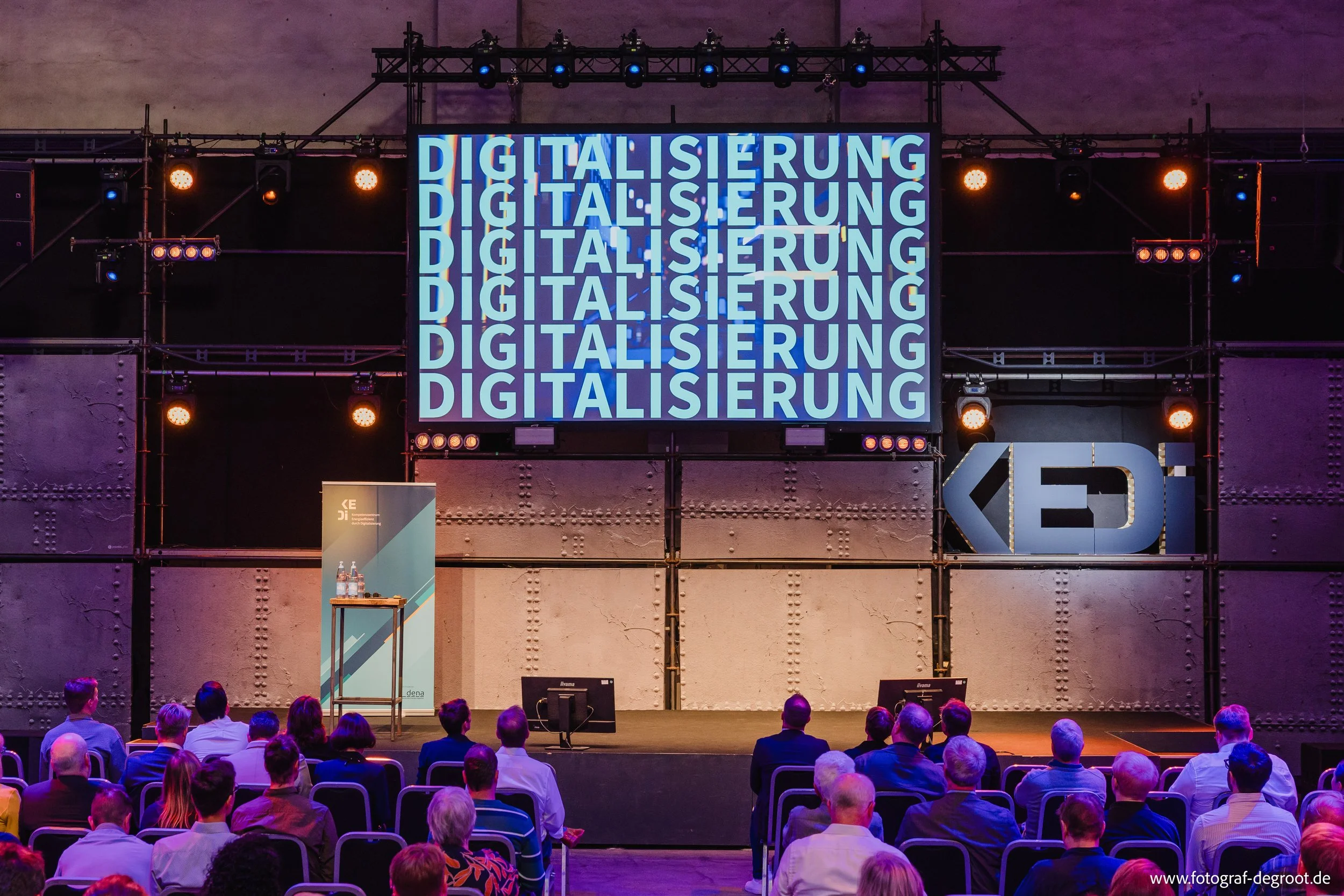 Bühne mit dem Wort 'DIGITALISIERUNG' in mehreren Reihen, umgeben von einem Publikum bei einer Konferenz.