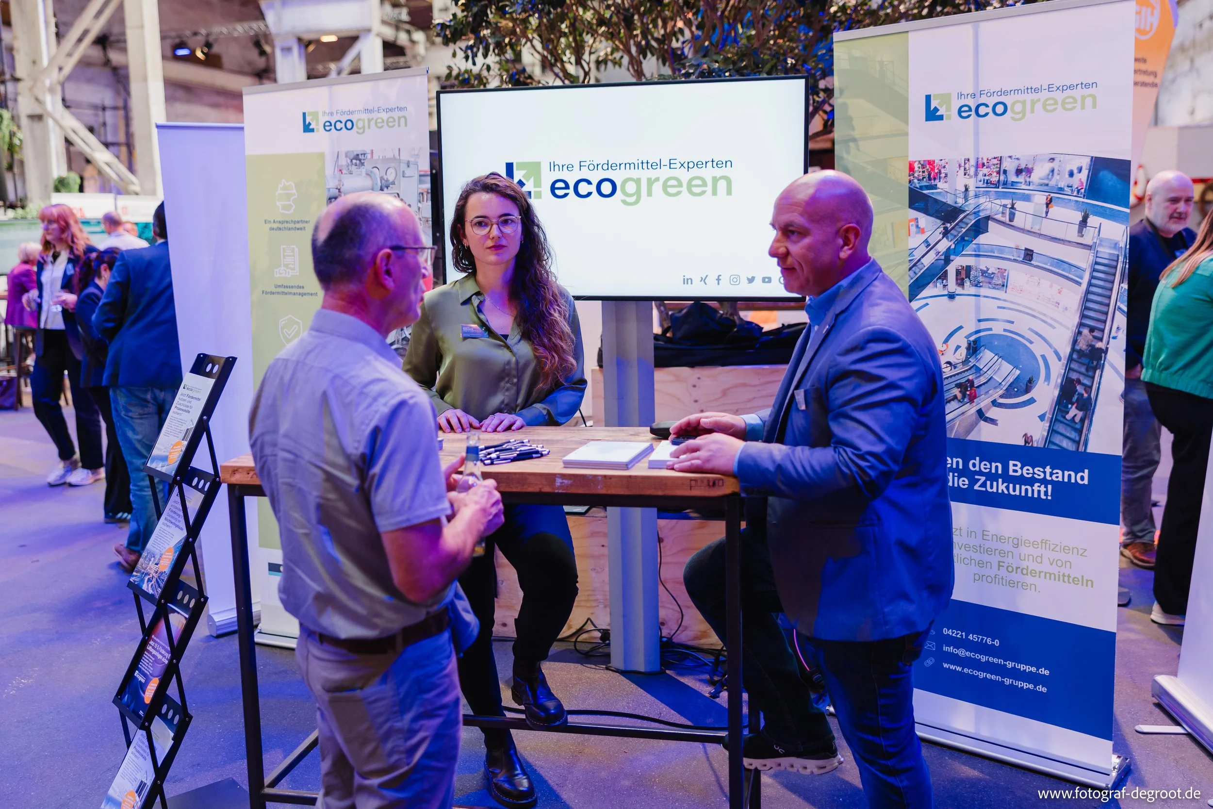 Menschen sprechen bei einer Messe an einem Stand, der von ecogreen vertreten wird, mit einem großen Bildschirm im Hintergrund, der das Logo und Informationen zeigt.
