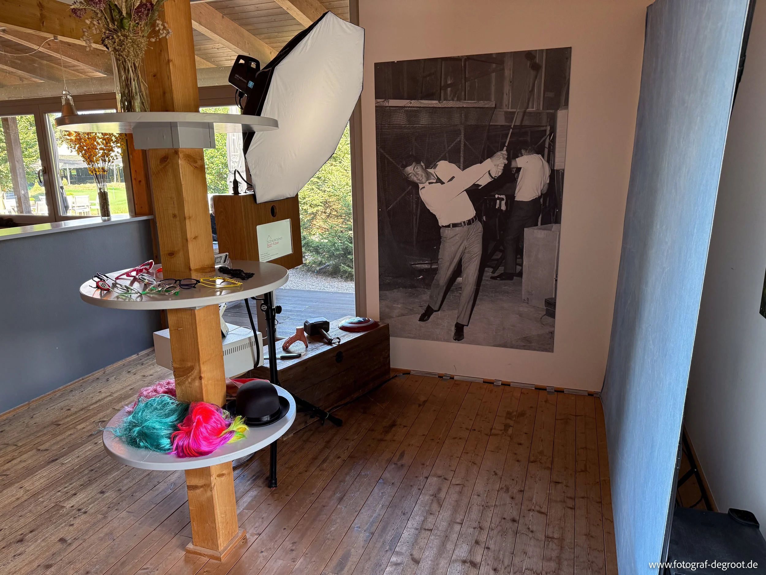 Ein Innenraum mit einem Holzfußboden, einer weißen Wand und einer großen schwarzen und weißen Fotografie an der Wand. Es gibt ein Stehpult mit bunten Perücken, Brillen und anderen Accessoires, sowie einem Filmlicht und anderem Equipment auf einem Tis