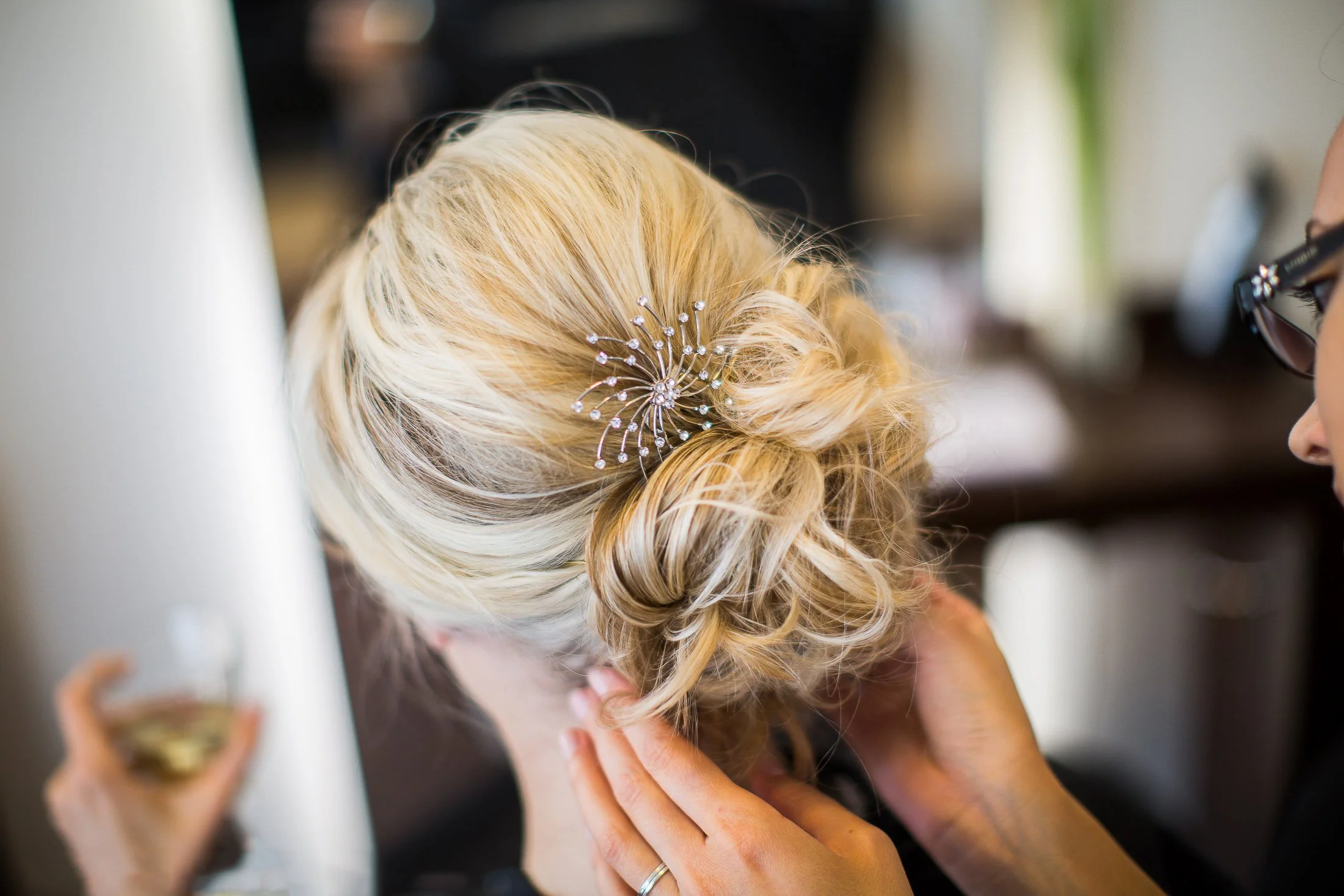 Nahaufnahme einer Frau mit blonden, zu einem lockeren Knoten gebundenen Haaren, verziert mit einem filigranen Haarspangen, während sie sich im Spiegel frisiert.
