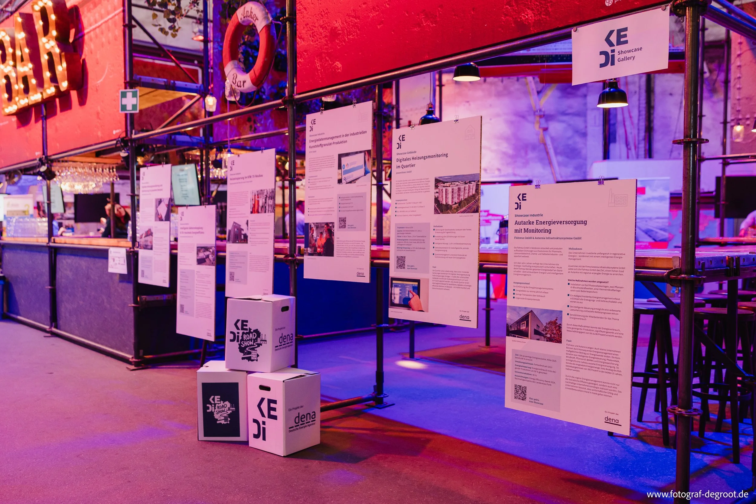Ausstellungsstand mit mehreren Informationsplakaten, die an einer Metallrahmenwand befestigt sind, in einem Raum mit bunten LED-Beleuchtung und Barbereich im Hintergrund.