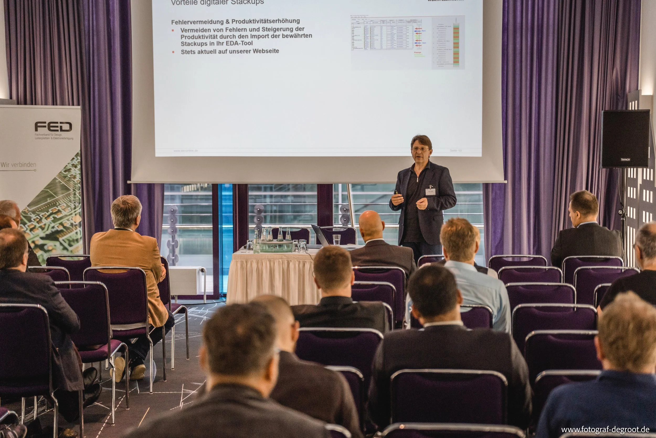 Präsentation in einem Konferenzraum mit einem Redner, der vor einem Publikum spricht. Eine Projektionsleinwand zeigt Folien über "digitale Stackups". Personen sitzen in Reihen und hören zu. Ein Tisch mit Getränken steht seitlich.
