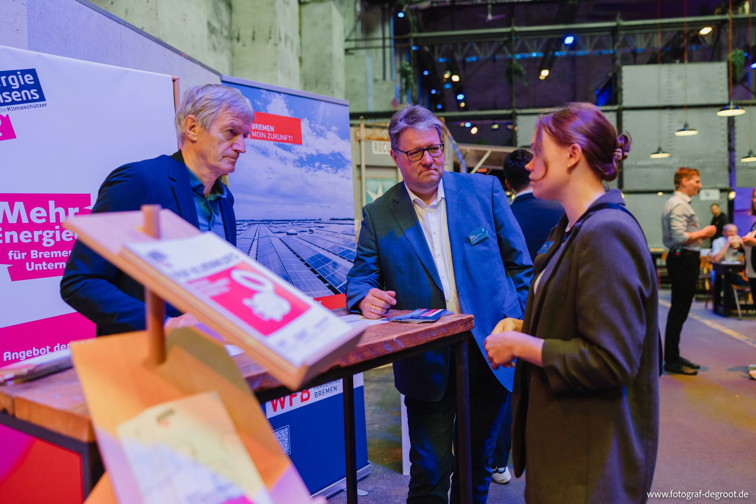 Drei Personen im Gespräch an einem Stand bei einer Veranstaltung. Der Stand bewirbt erneuerbare Energien für Bremen, erkennbar an den Plakaten mit Solarzellen und Energieförderung.