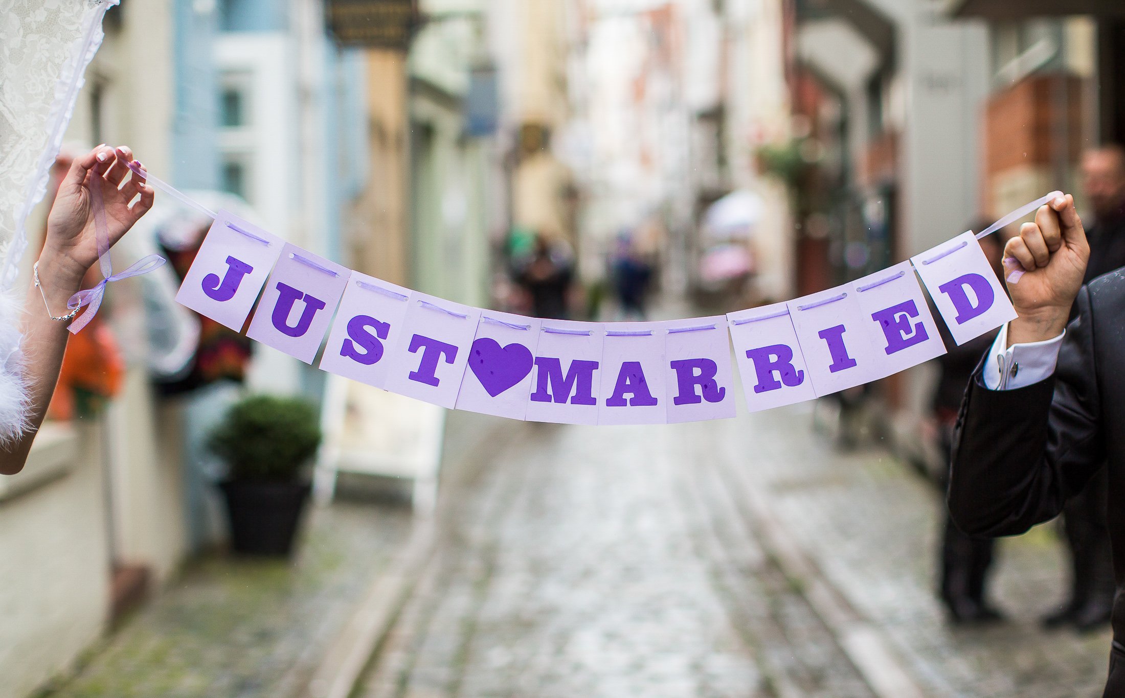 Menschen halten ein Banners mit der Aufschrift "JUS♥MARRIED" während einer Feier auf einer belebten Straße, vermutlich eine Hochzeits- oder Verlobungsfeier.