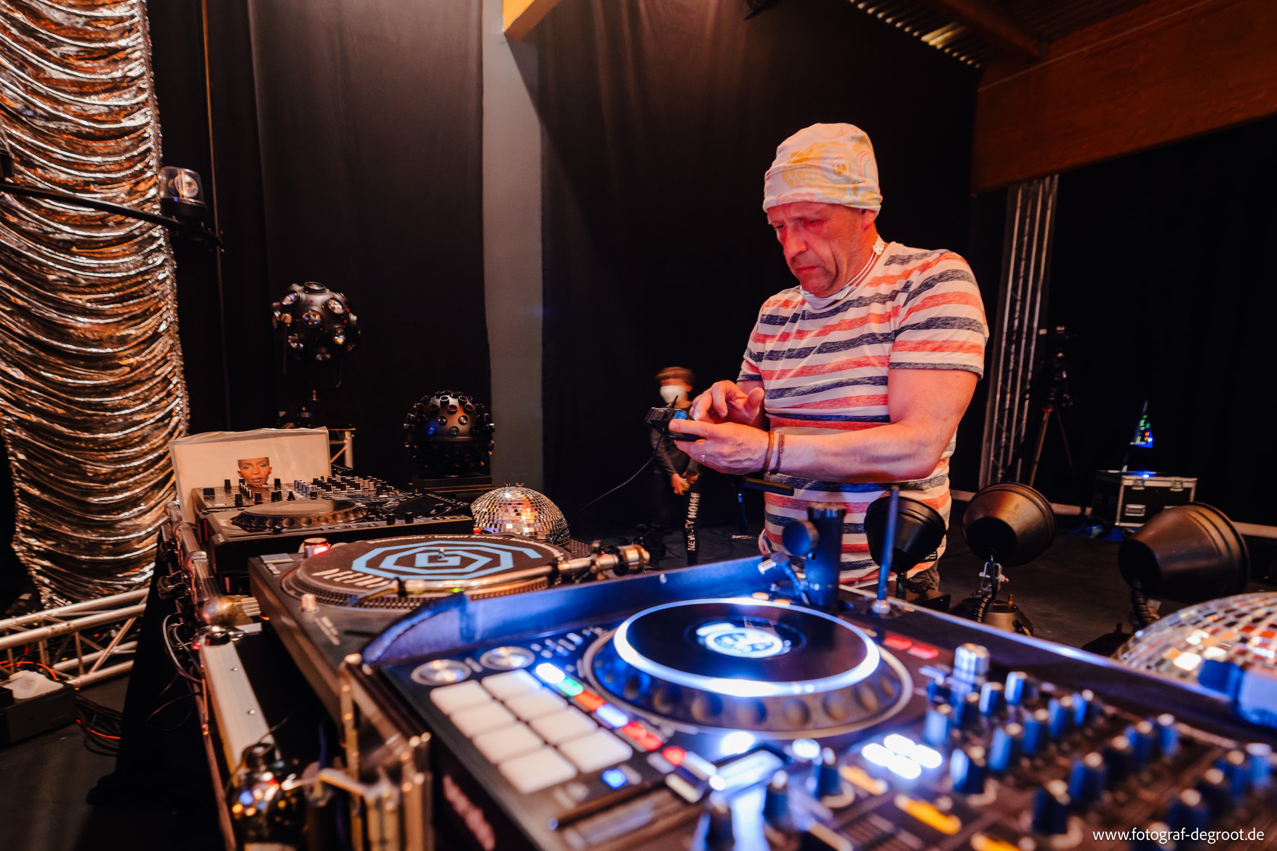 DJ bei der Arbeit mit Mischpult und Turntable, trägt ein gestreiftes T-Shirt und eine Mütze, Bühne mit Lichtequipment im Hintergrund.