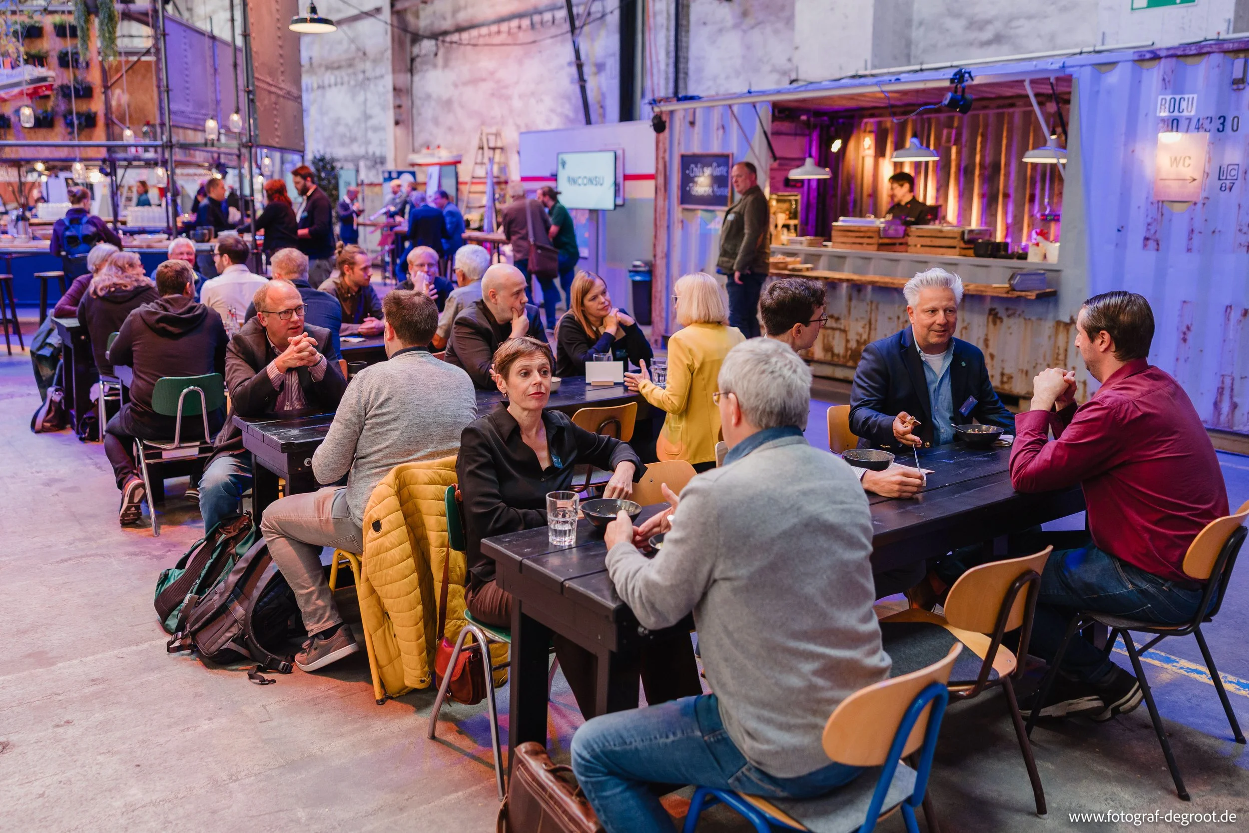 Menschen sitzen an Tischen in einem Veranstaltungsraum mit Industriecharme, trinken und sprechen. Im Hintergrund gibt es eine Bar und einen Essensstand, dekoriert mit Lichterketten und Neon-Lichtern.