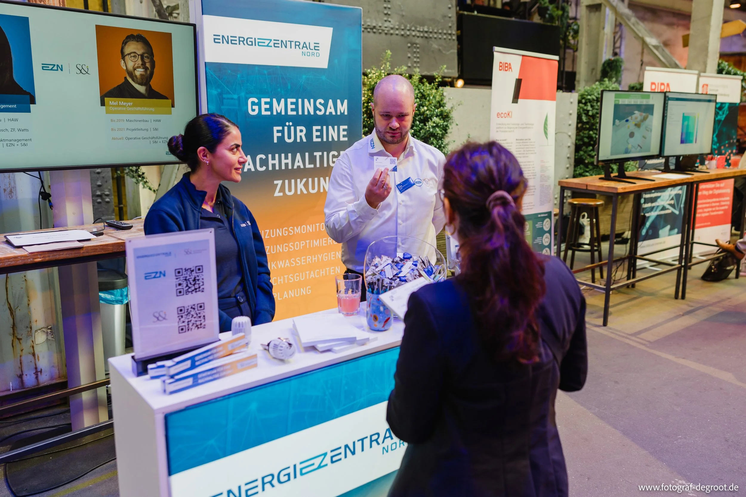 Menschen bei einer Messe oder einem Event, Gespräche am Stand für eine nachhaltige Zukunft, mit Infomaterial und Bildschirmen im Hintergrund.