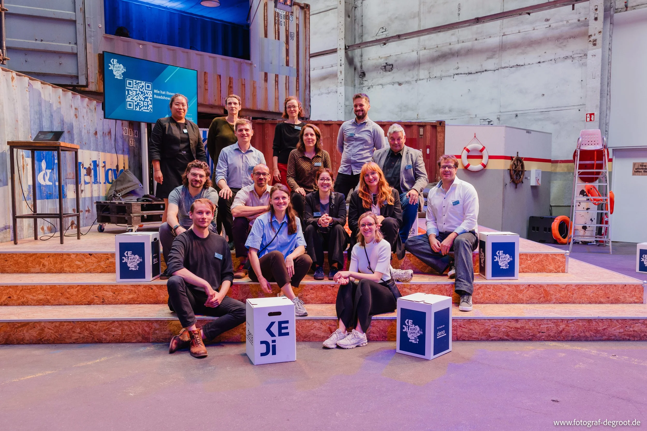 Gruppe von circa 15 Menschen posiert auf einer kleinen Bühne in einer Industriehalle. Im Hintergrund sind Container, ein Bildschirm mit QR-Code und Life Ringe sichtbar.