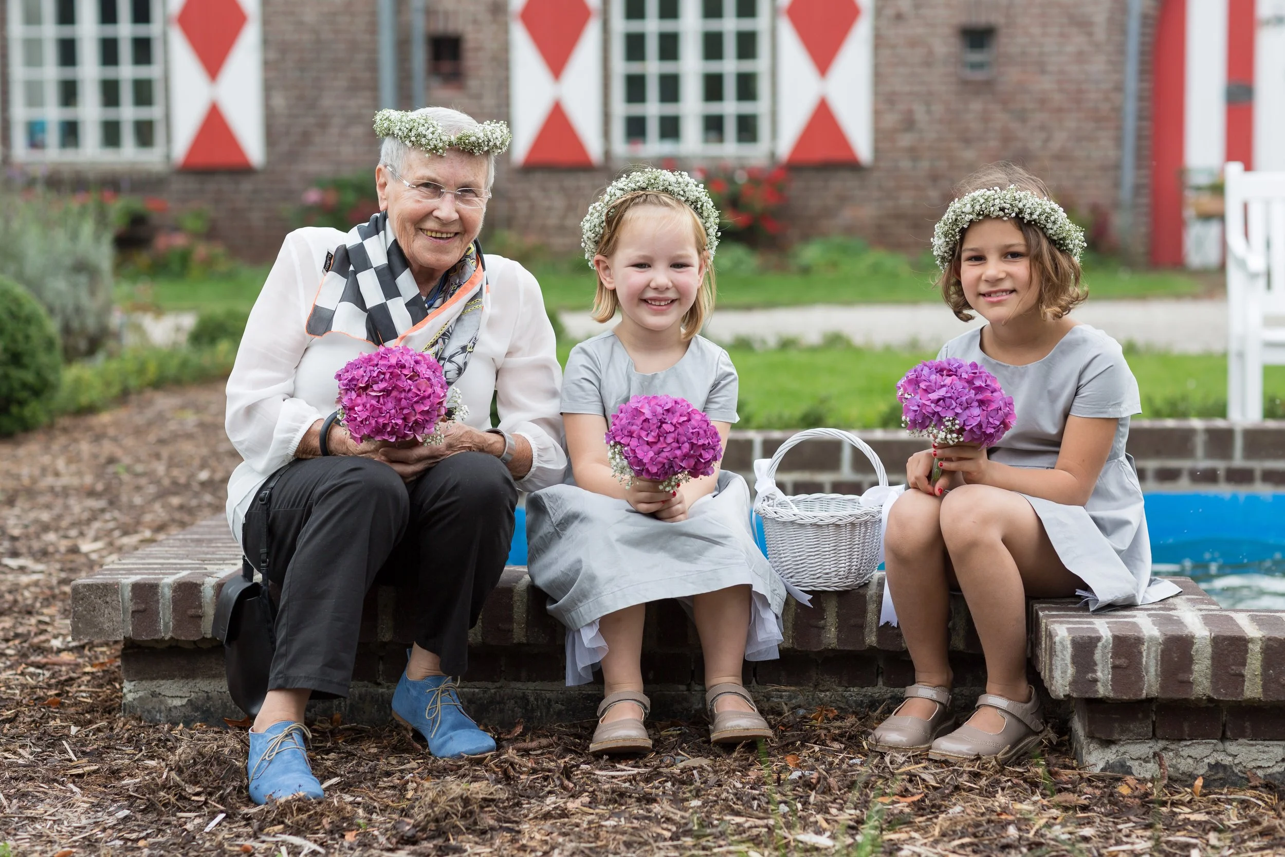 Drei Kinder und eine ältere Frau sitzen auf einer Brücke, alle tragen Blumenkränze und halten Blumensträuße, vor einem roten Backsteingebäude mit weißen Fenstern.