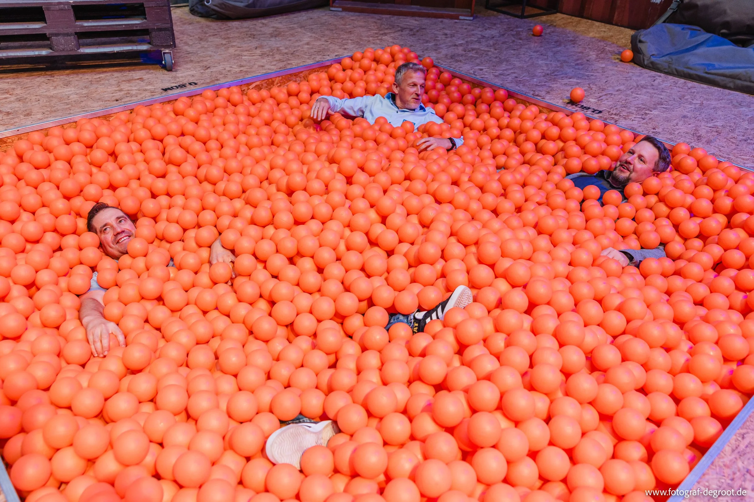 Drei Männer liegen in einer Ballgrube aus orangefarbenen Bällen, die auf einer Bühne im Innenraum eines Gebäudes verteilt sind.