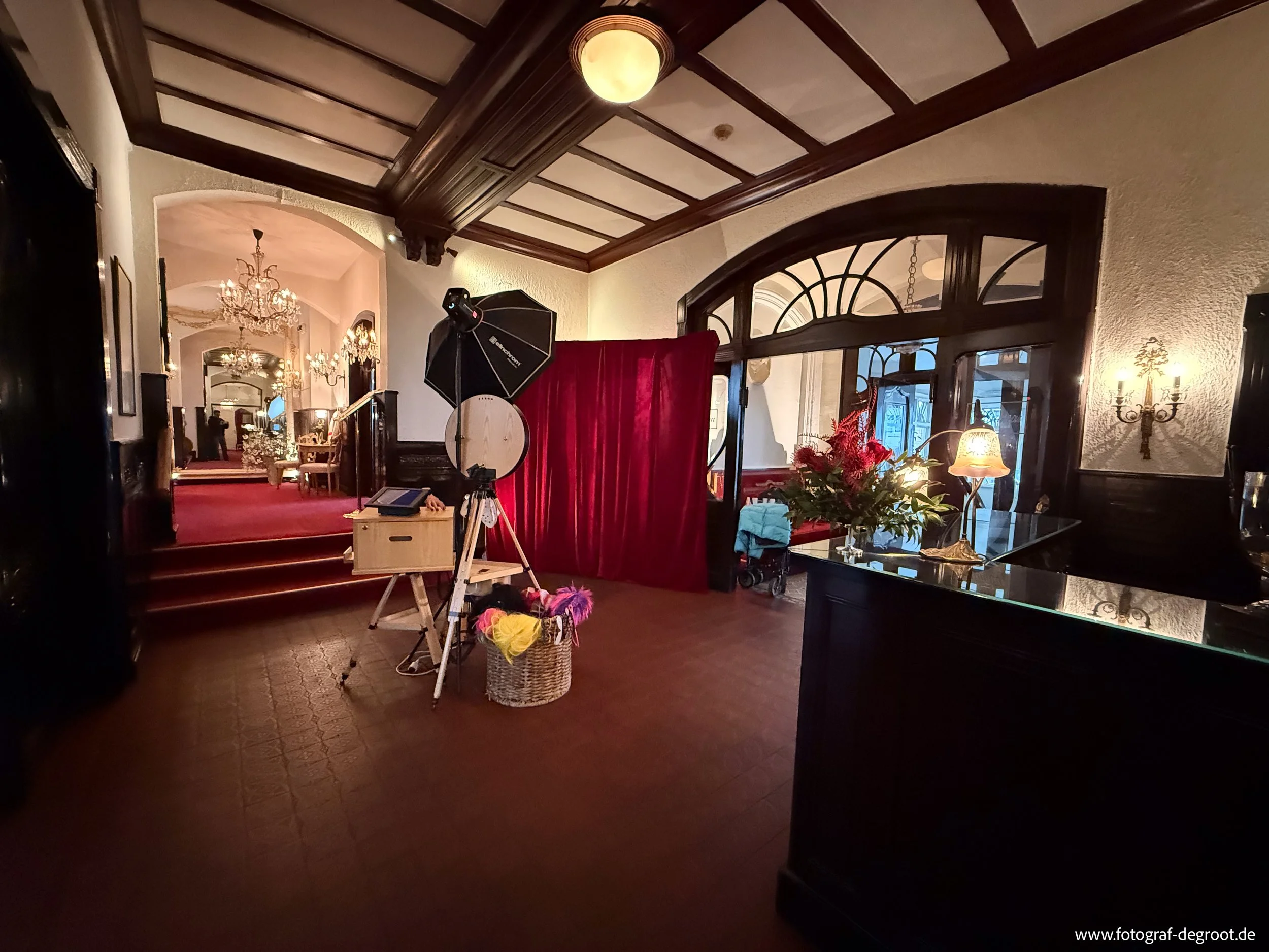 Innenaufnahme eines eleganten Hotel- oder Restaurantbereichs mit dunklem Holzdekor, einem roten Vorhang, Blumenstrauß, Fototechnik, und einem Treppenaufgang mit Spiegel und Kronleuchtern im Hintergrund.