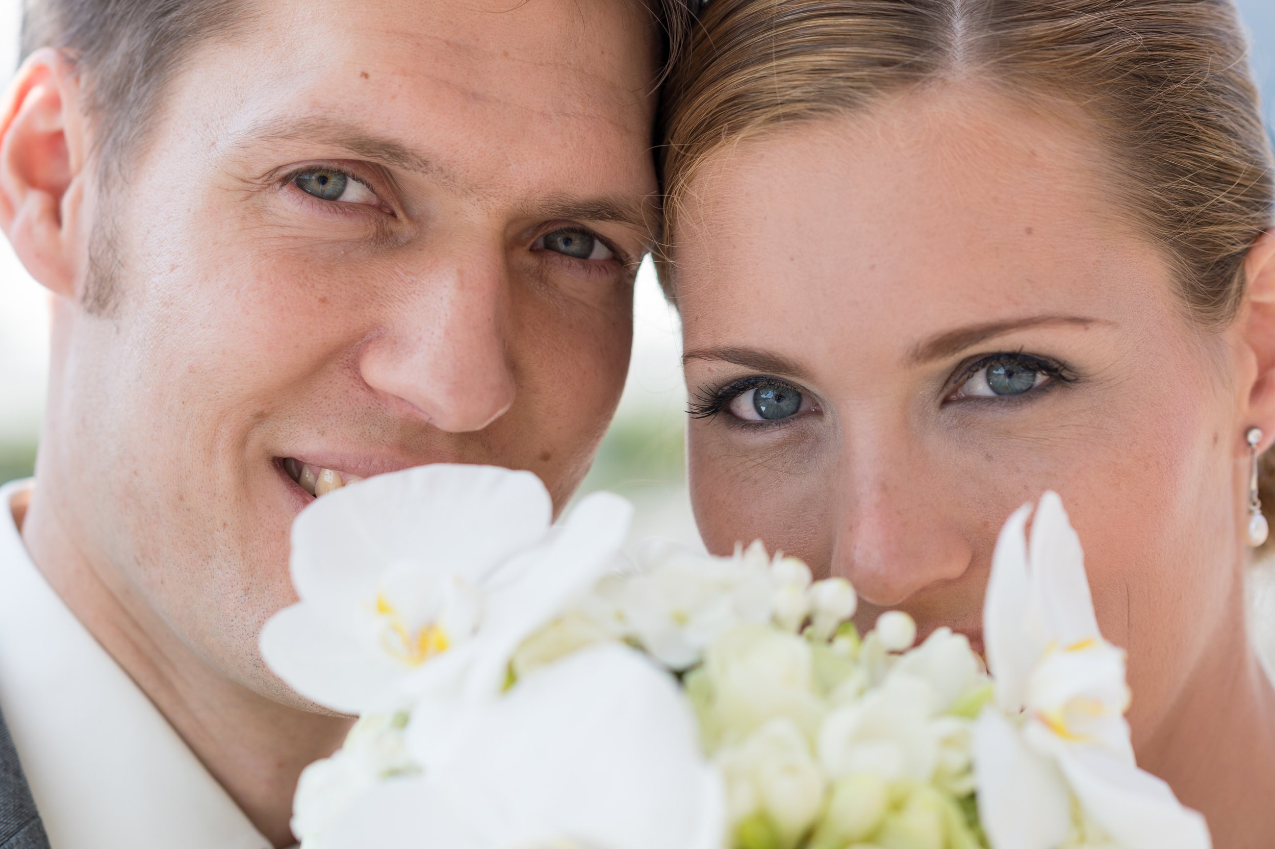 Ein Mann und eine Frau stehen ganz nah beieinander, schauen in die Kamera und lächeln. Im Vordergrund sind weiße Blumen, möglicherweise eine Hochzeit.