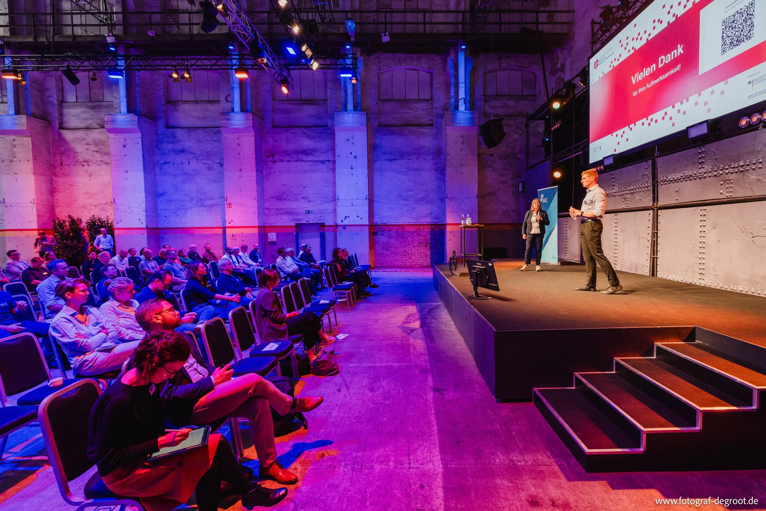 Eine Konferenz mit einem Präsentationsbildschirm, auf dem steht 'Vielen Dank für Ihre Aufmerksamkeit'. Es gibt zwei Sprecher auf der Bühne, während das Publikum im Saal sitzt und zuhört.
