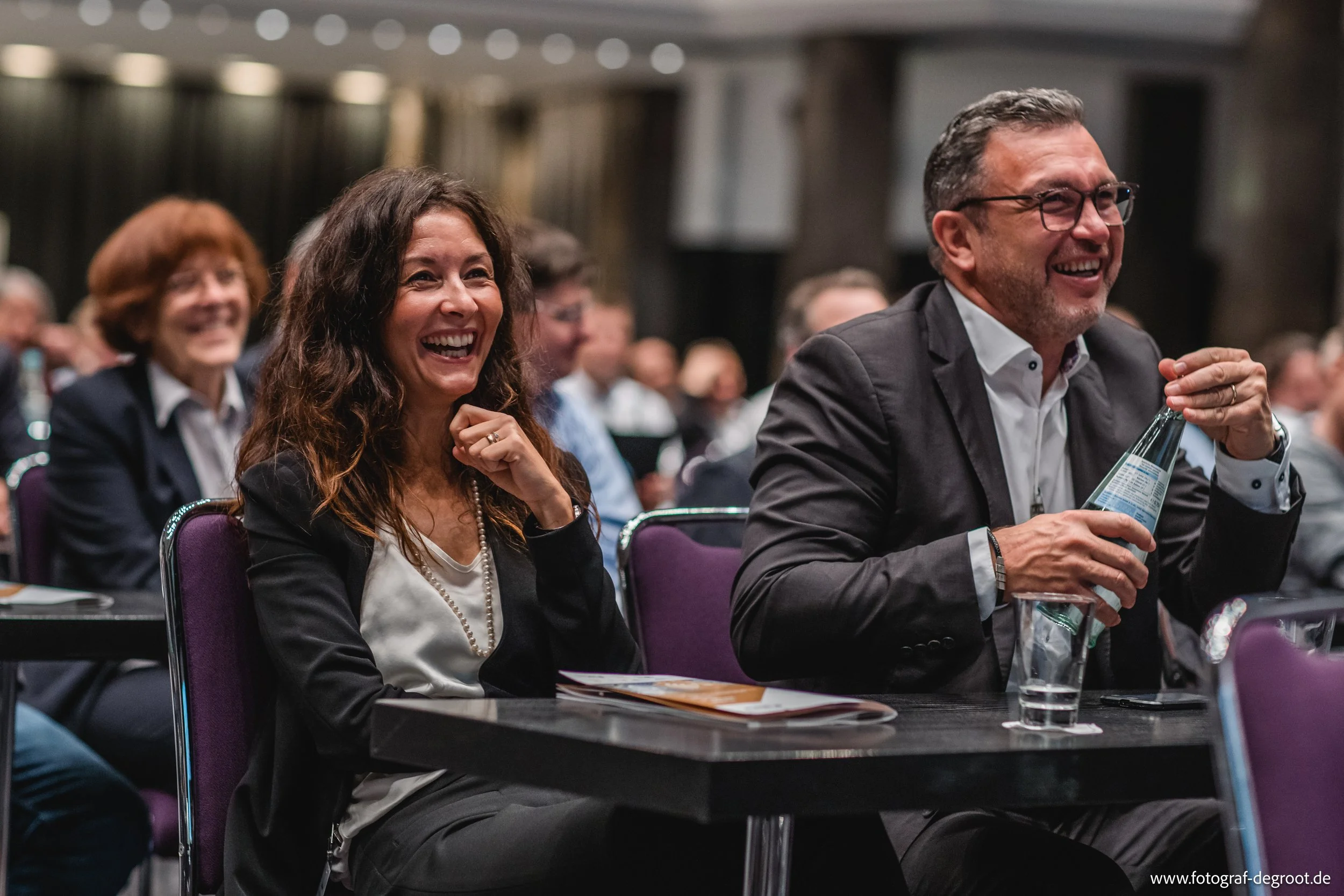 Menschen lächeln und lachen bei einem Event oder einer Konferenz, während sie an Tischen sitzen.