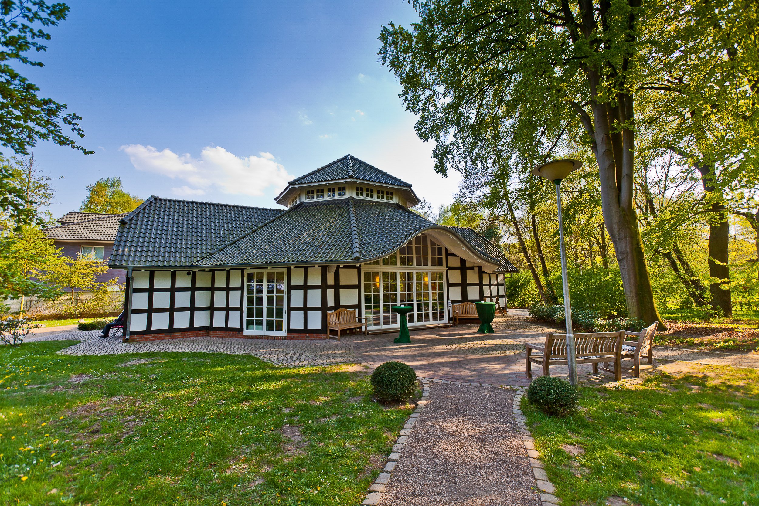 Ein gepflegtes Haus im Garten, umgeben von Bäumen und Sitzbänken, bei sonnigem Himmel.